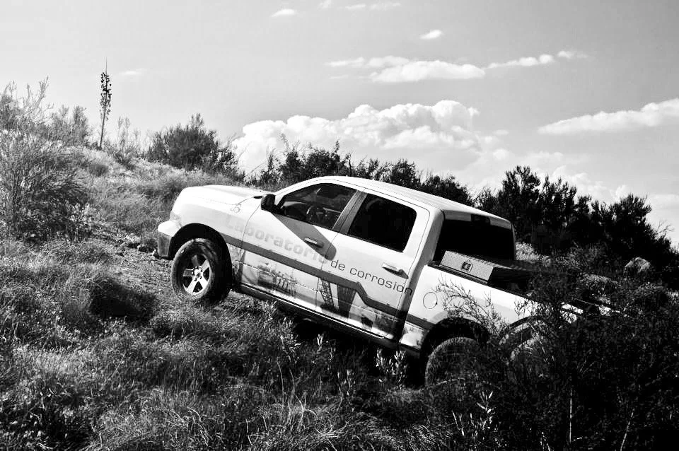 A white pickup truck is stuck on a grassy hillside with bushes, with a cloudy sky overhead.