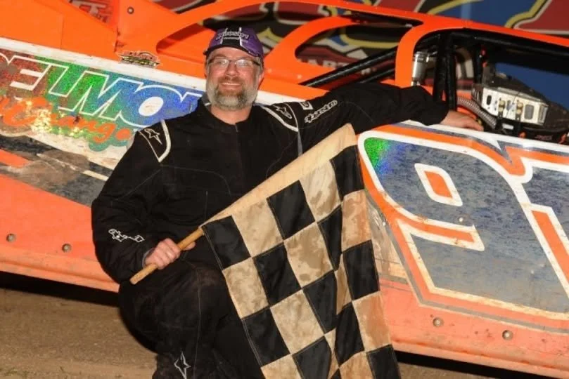 A race car driver with a beard, in racing gear, holding a checkered flag, crouching next to his orange race car at a dirt track.