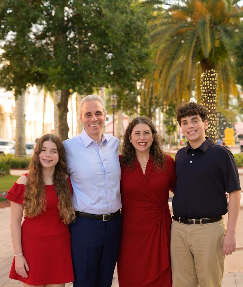 Family of four, two adults and two children, standing outside on a city street with palm trees, smiling and posing for the photo.
