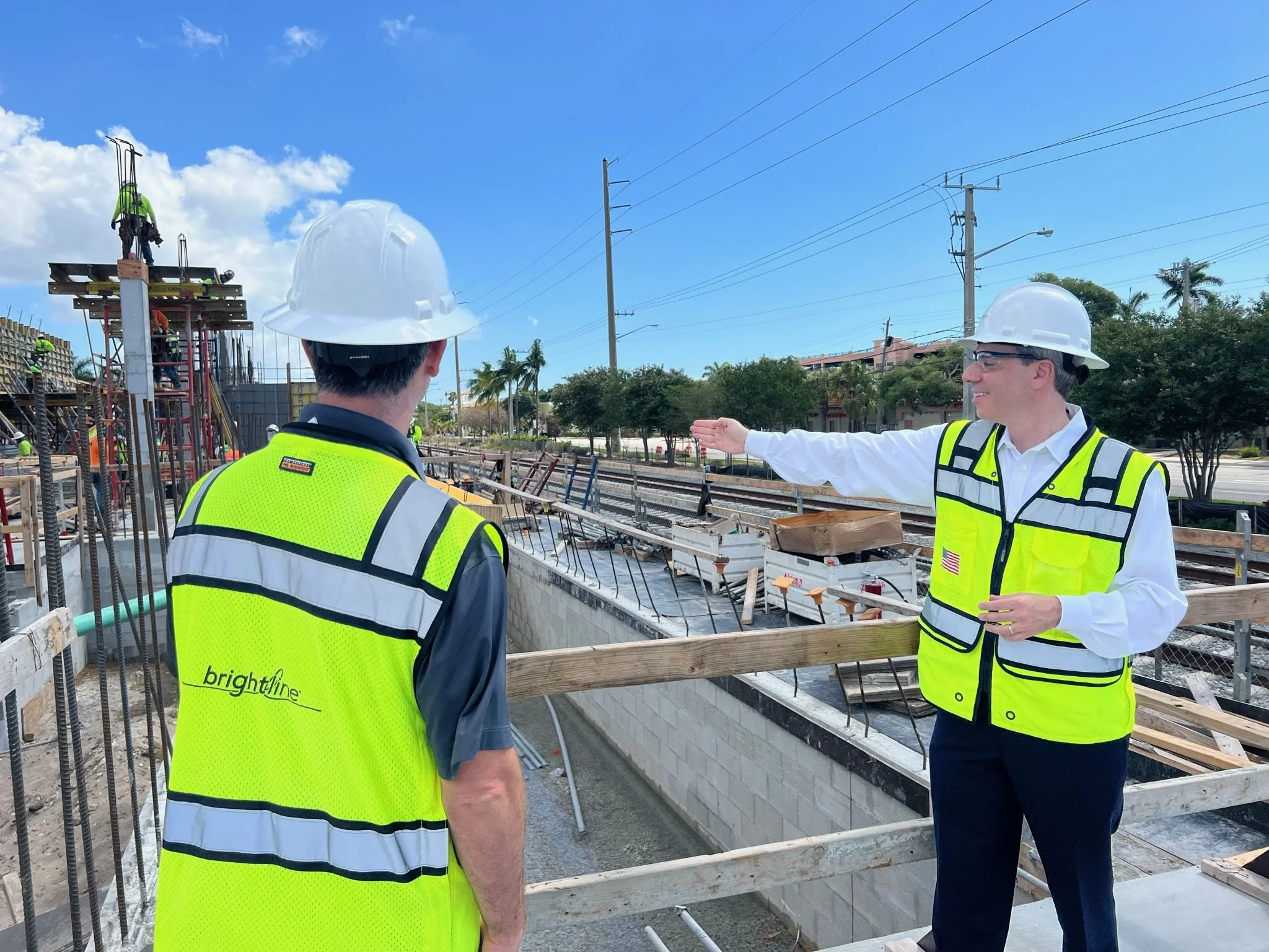 Two construction workers in safety vests and hard hats standing on a construction site near train tracks, with one person pointing and the other listening attentively.