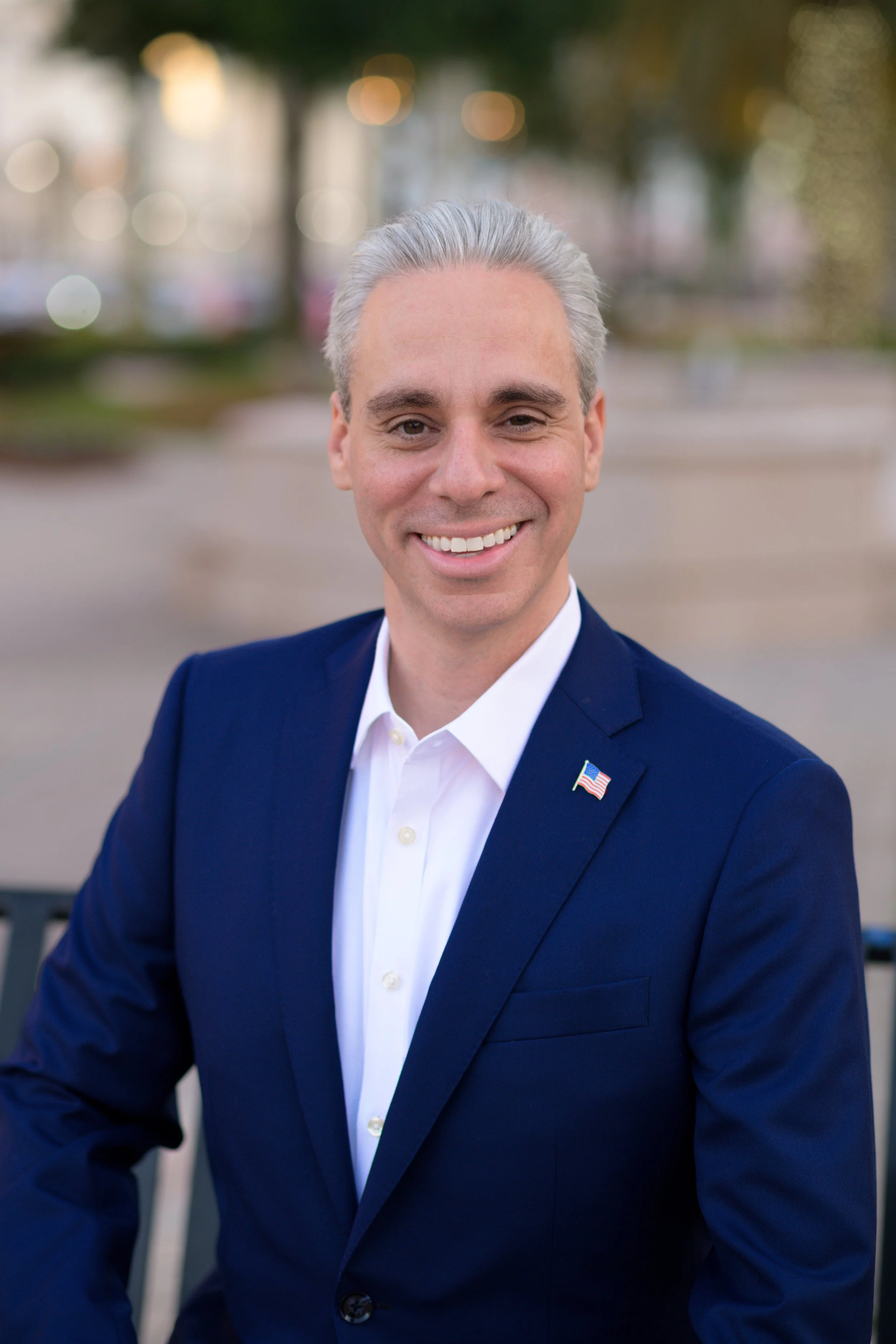 A man with gray hair, dressed in a blue suit and white shirt, smiling outdoors with blurred trees and lights in the background.