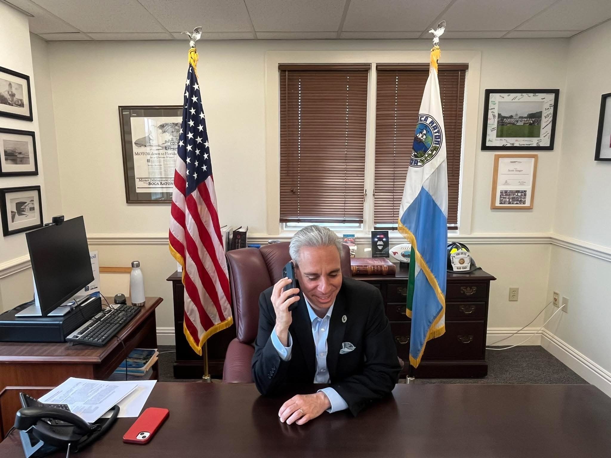 A man in a suit sitting at a desk, talking on a landline phone, with two flags behind him in an office. The flags are the United States flag and another flag, with a window with blinds, artwork, and office supplies visible.