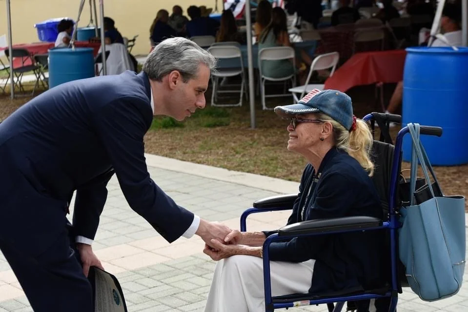 A man in a suit is holding hands and shaking hands with an elderly woman who is sitting in a wheelchair, wearing a cap and glasses at an outdoor event.