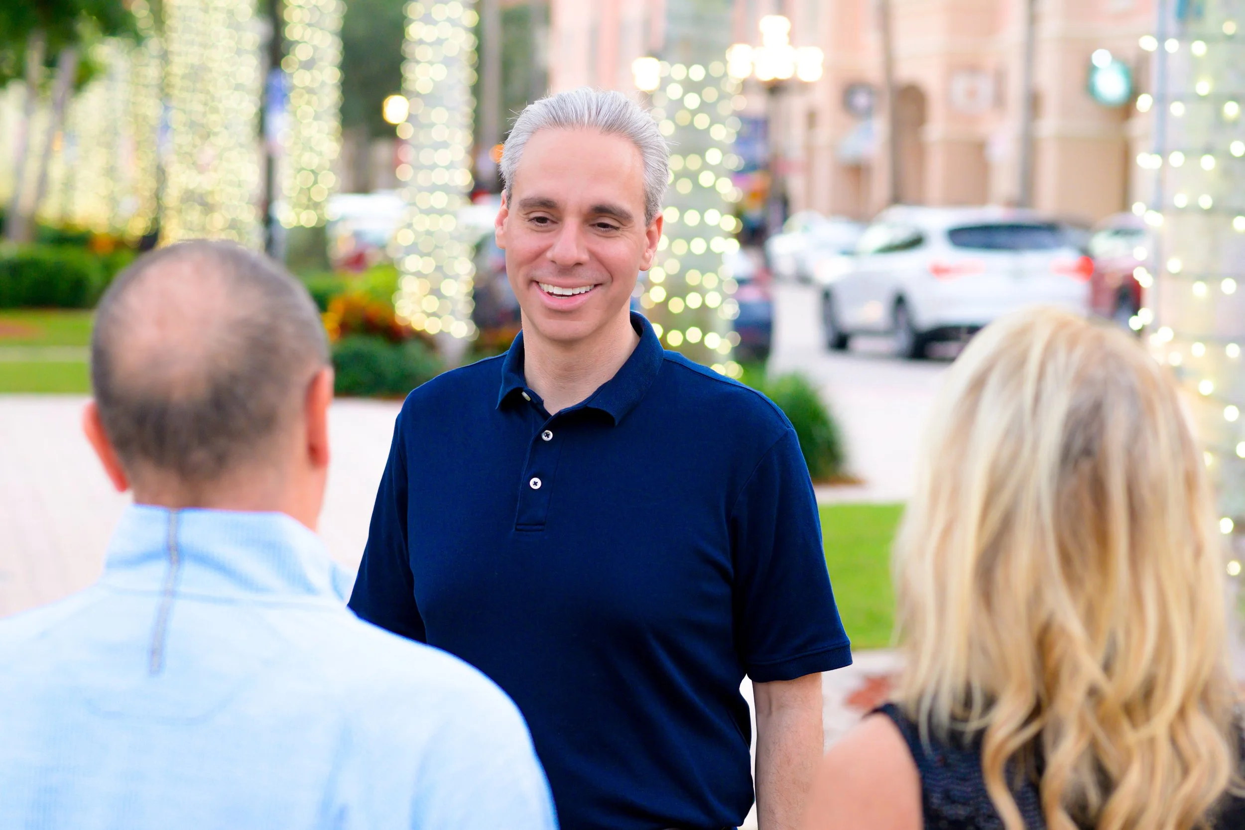 A man with gray hair smiling and talking to a couple outdoors on a decorated street with lights.
