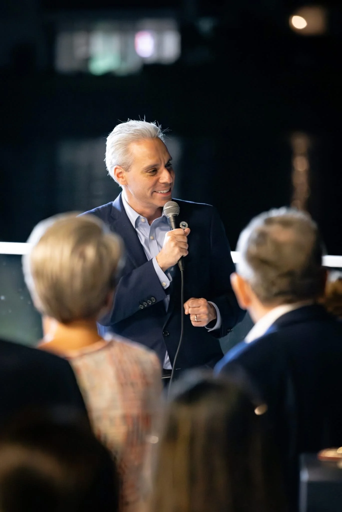 A man with gray hair wearing a dark suit jacket speaking into a microphone in front of an audience.