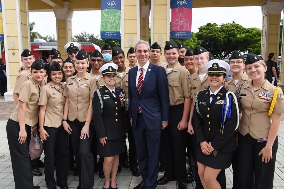 Group of young people in military uniforms posing with a man in a suit in front of a pavilion with banners.