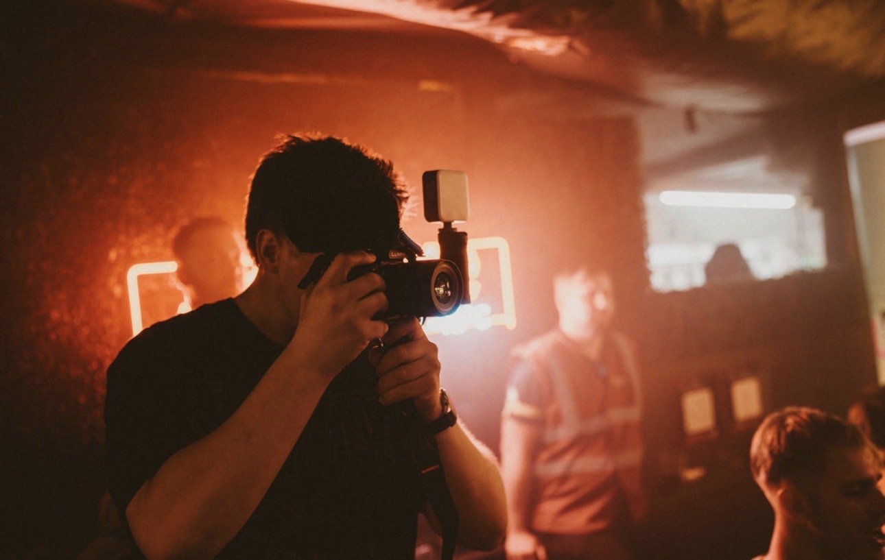 A person holding a camera with a flash attachment, taking a picture in a dimly lit room with orange-red neon lights and other people in the background.