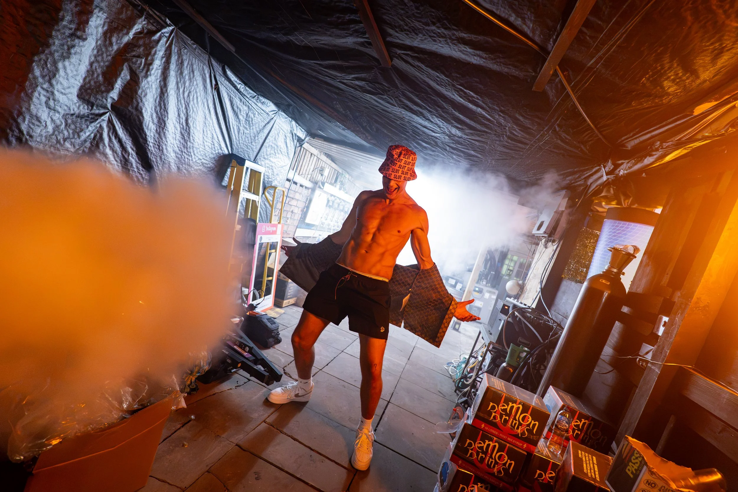 A young shirtless man wearing a bucket hat with the word 'SLUT' repeated all over, shorts, white socks, and sneakers, standing in a dimly lit room with black plastic sheeting on the walls. He appears to be dancing or posing, with a wide smile on his face. The room has various electronic equipment, boxes, and a smoky or foggy atmosphere. There are bright lights illuminating the scene from behind.