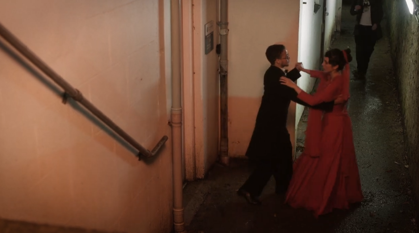 A couple dancing closely in a narrow alley at night, with a woman in a red dress and a man in a black suit. An additional person is visible in the background.