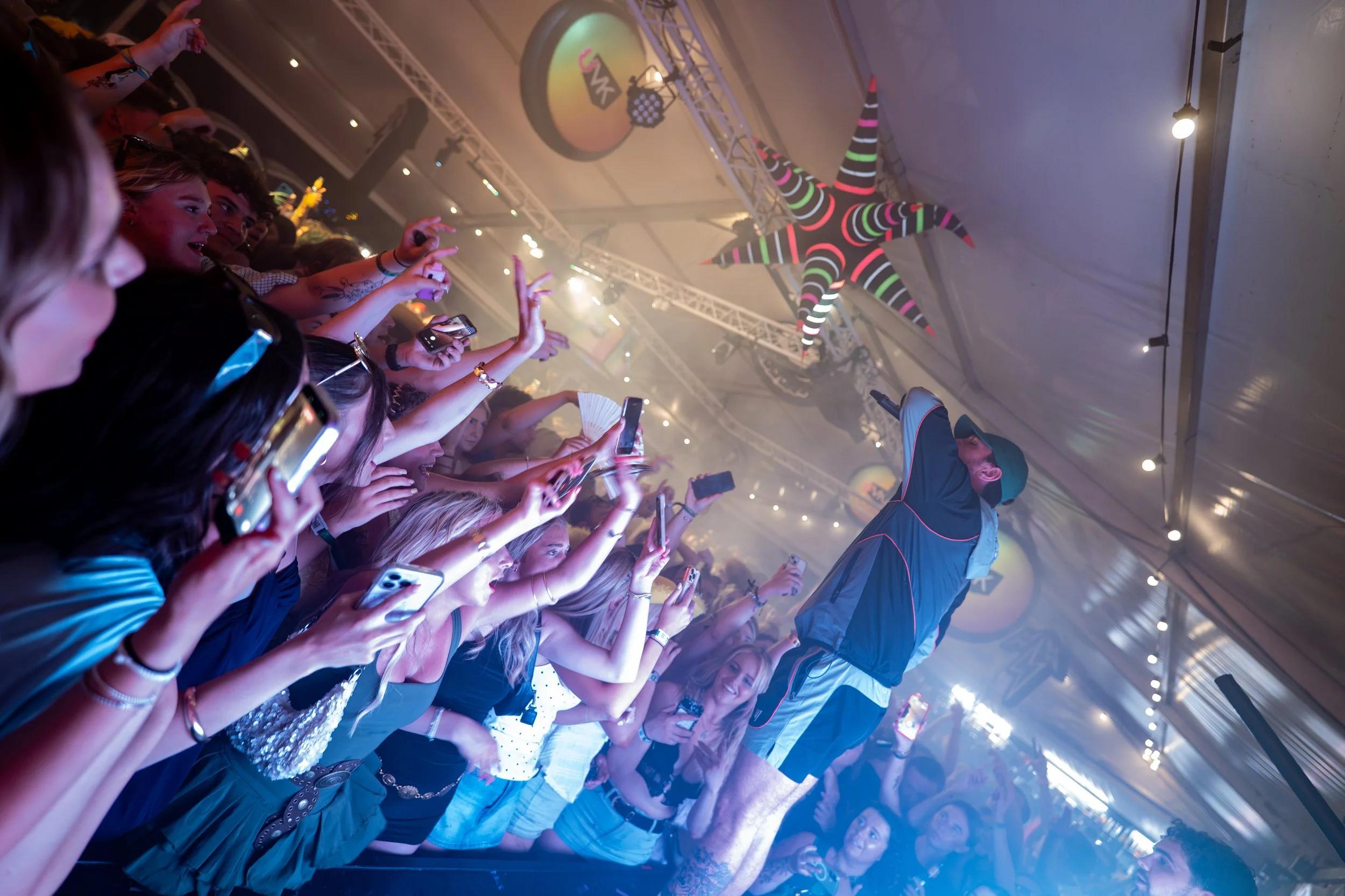 Crowd of people at a concert holding up phones, with a large star-shaped inflatable decoration hanging from the ceiling, and stage lighting.