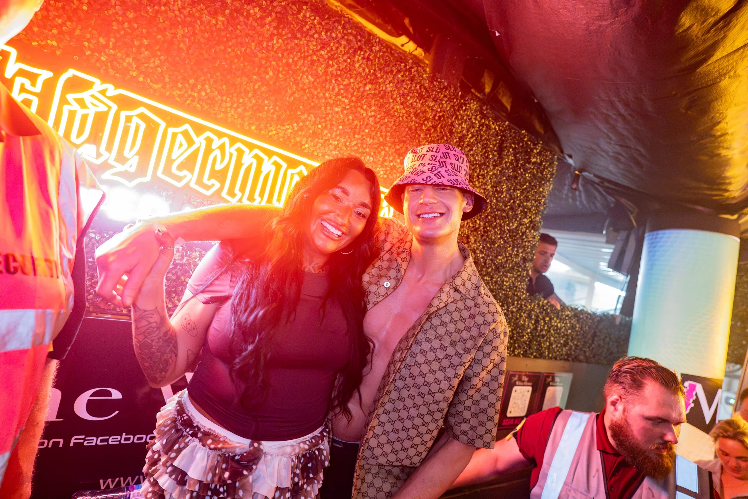 Two people smiling, posing for a photo at a lively event, with neon signs and festive lighting in the background.