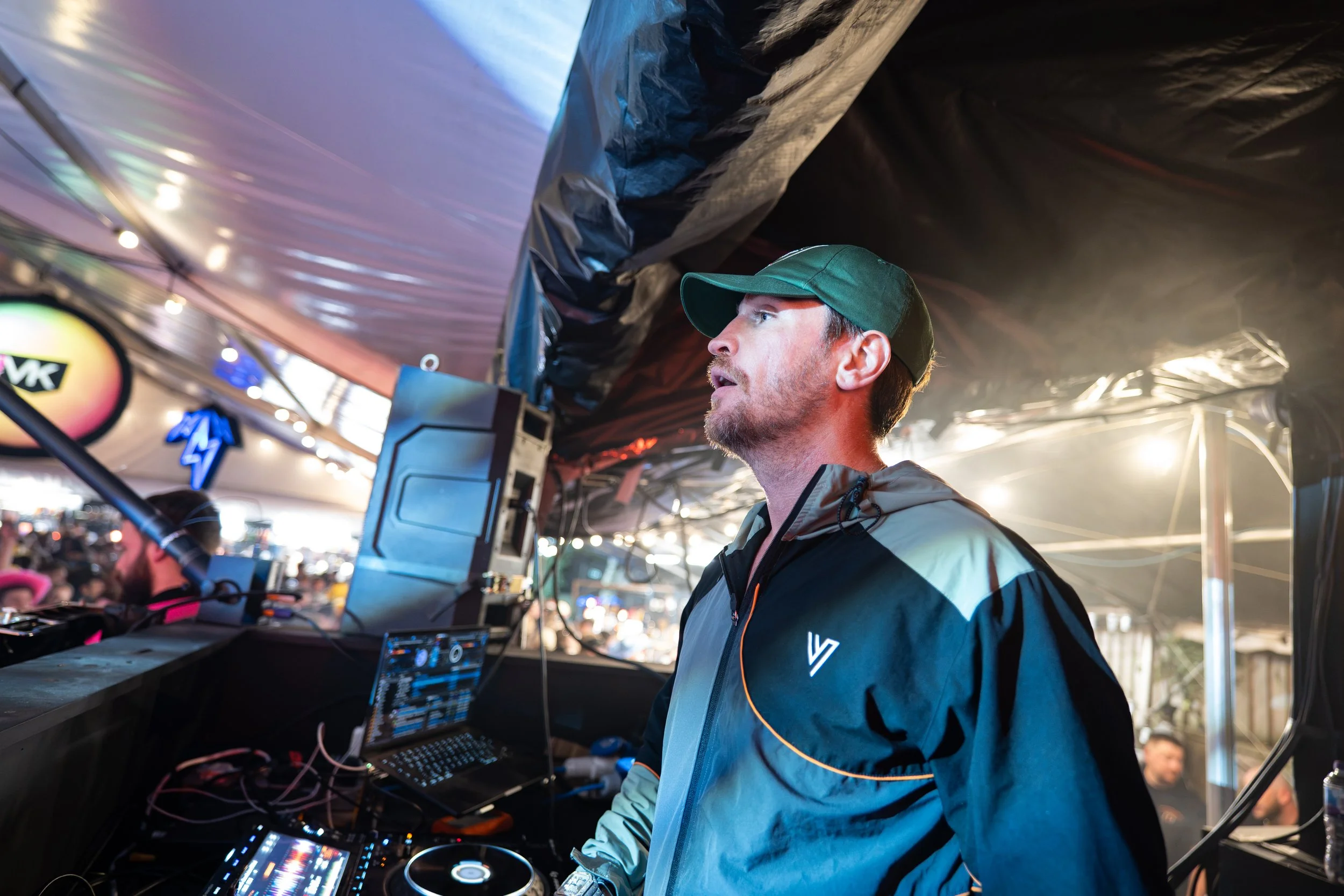 A man wearing a green cap and a jacket is DJing at an event, surrounded by equipment, with a crowd in the background under a tent with colorful lights.