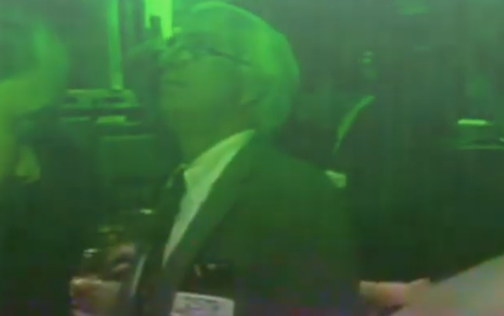 A person with blonde hair in a ponytail, wearing a dark manager shirt and in a green-tinted environment, possibly a security camera or night vision setting.