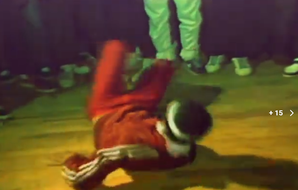 Person in red pants and black hat falling or performing a breakdance move on the floor with other people standing around, some in sneakers. The scene is indoors with dim lighting.