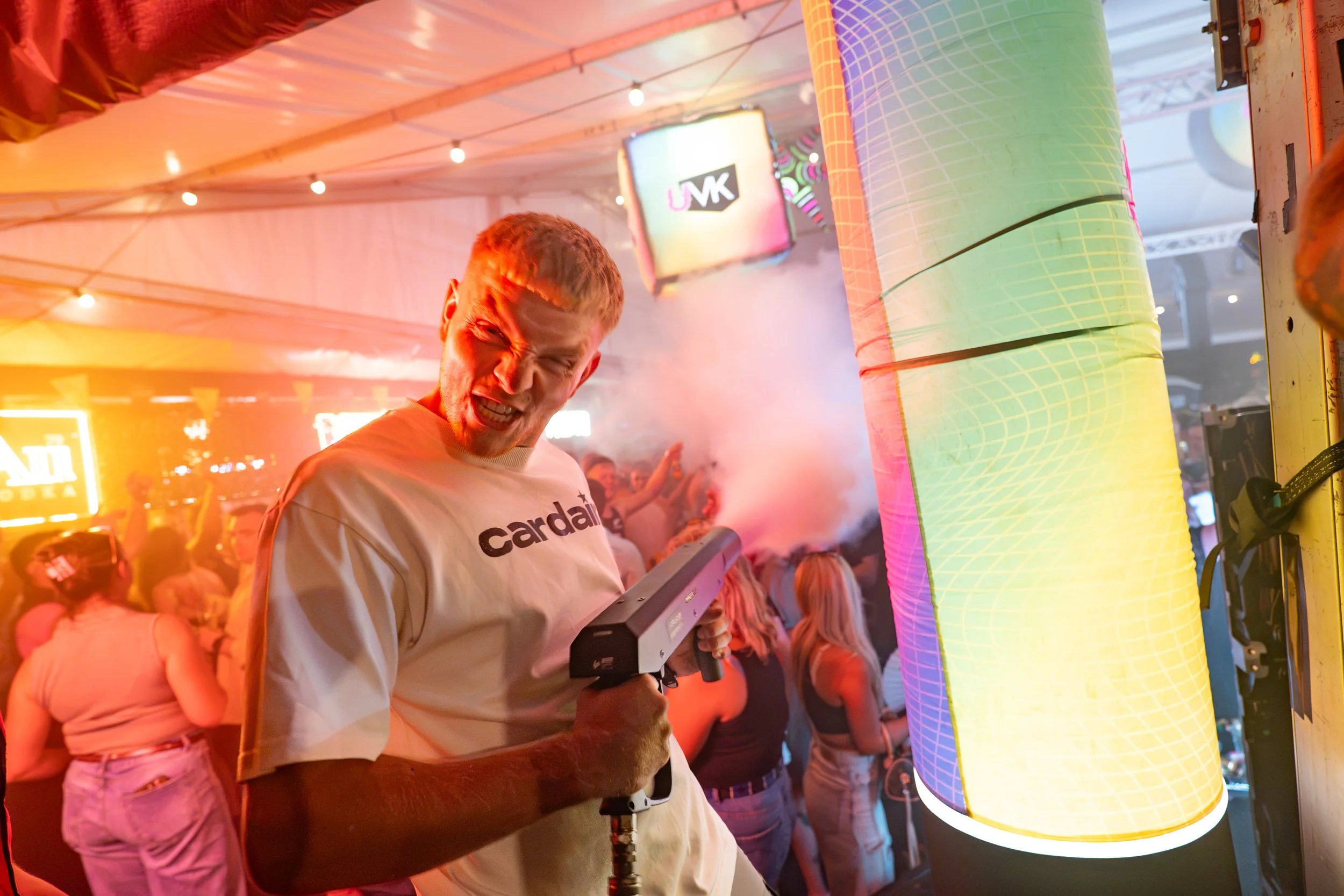A man with short blonde hair wearing a white shirt with the word 'Cardinal' on it is holding a gaming controller in a crowded indoor setting with colorful lighting, neon signs, and large inflatable structures.