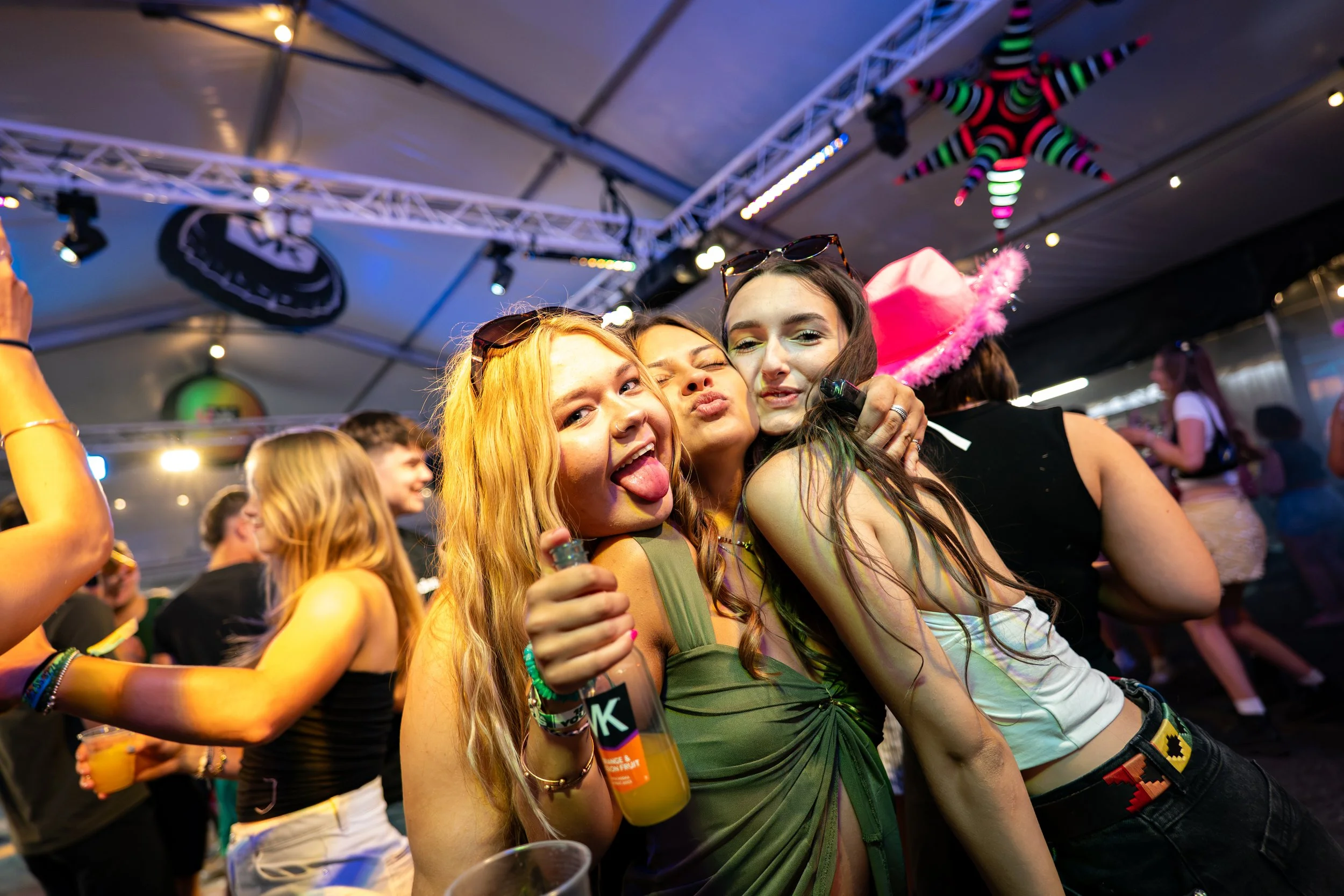 Group of young women partying at a lively indoor event, some wearing sunglasses and a pink carnival hat with a fluffy pom-pom, with colorful star decorations hanging from the ceiling in the background.