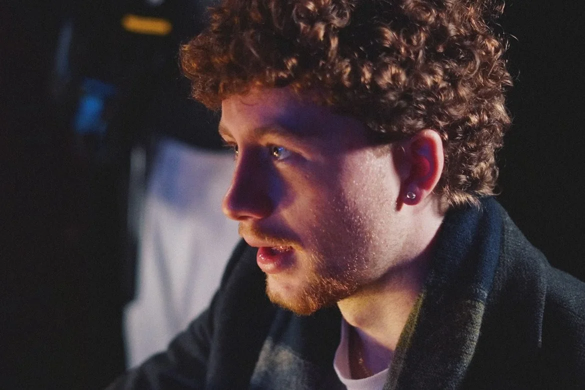 A young man with curly hair and a beard looking intently at something in front of him, with a focused expression, in a dimly lit environment.