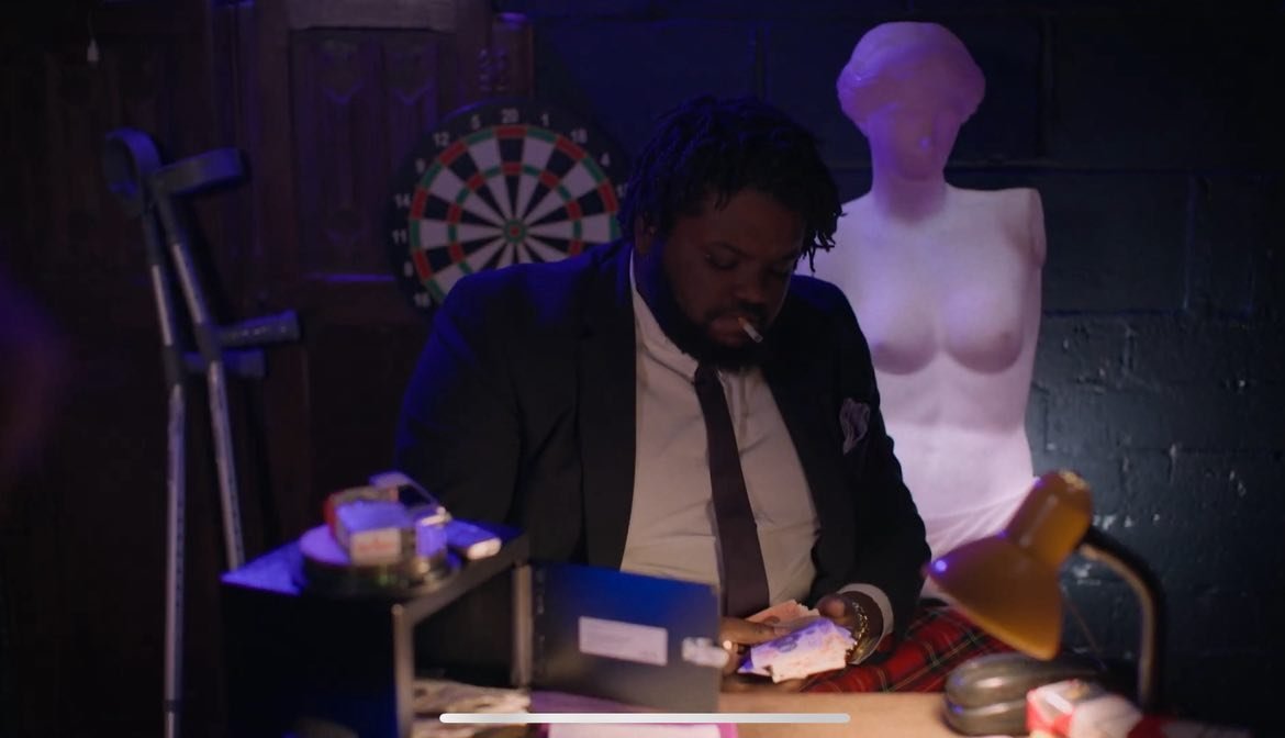 Man in suit sitting at a desk with crutches nearby, using a phone, with a dartboard on the wall and a female mannequin in the background.