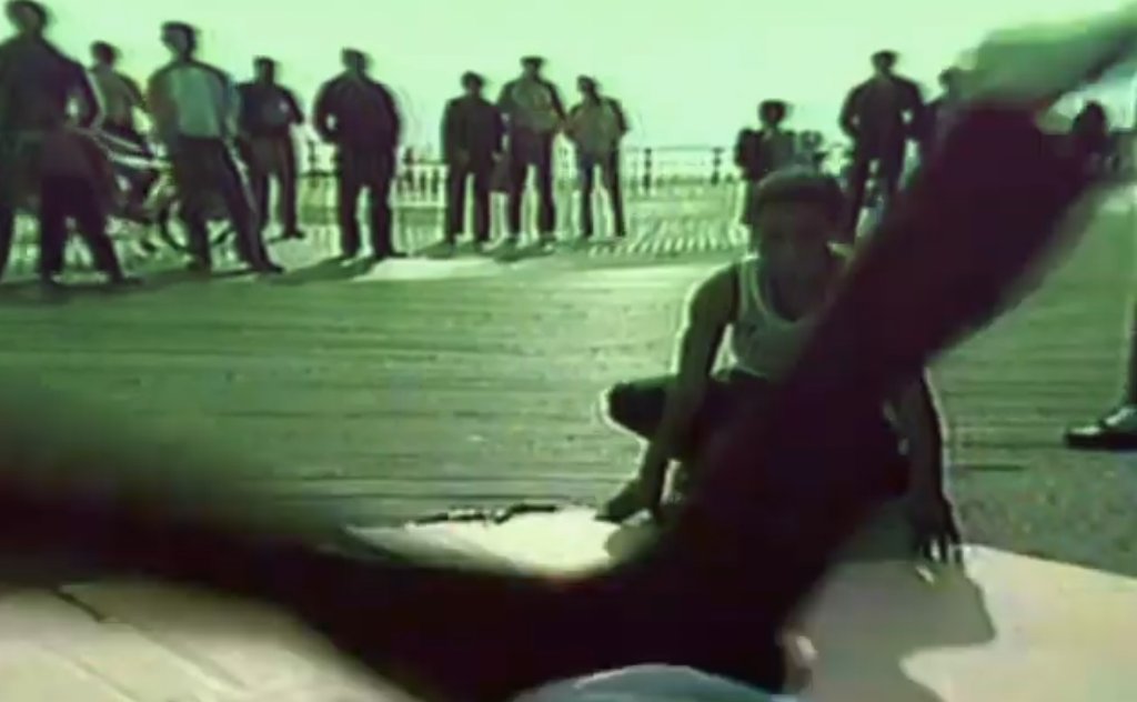 A young person in a white tank top sitting on a wooden deck, with a group of people standing in the background. The scene appears to be outdoors on a sunny day.