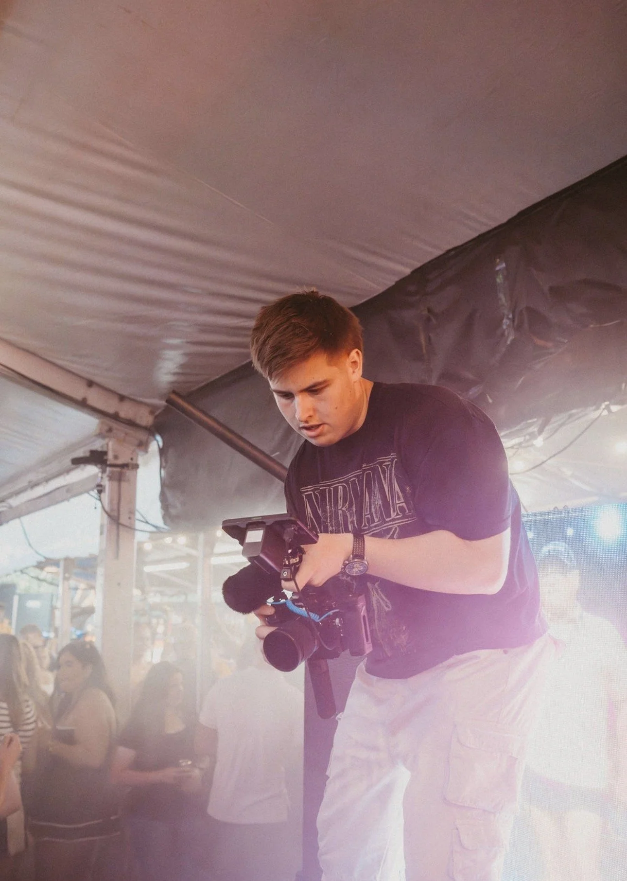 Young man with brown hair wearing a Nirvana t-shirt holding a camera inside a tent at a crowded event.