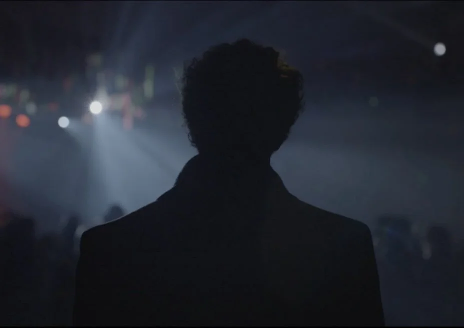 Silhouette of a person with curly hair in a dark room with stage lights in the background.