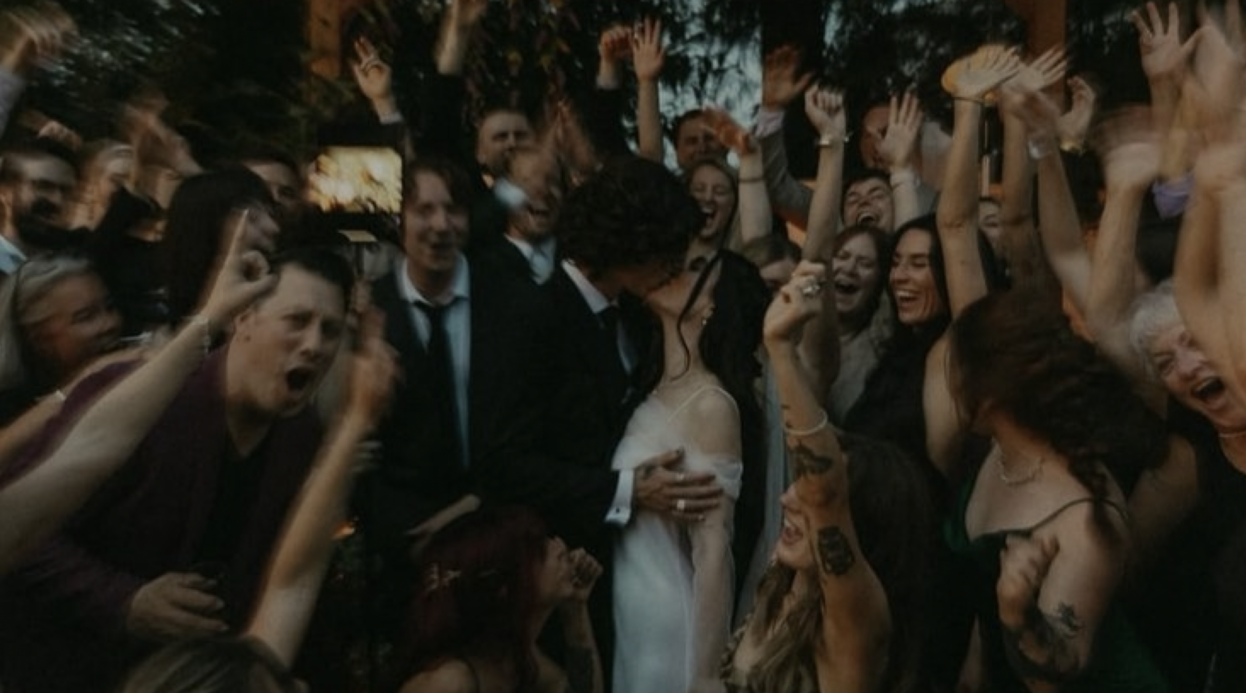 A group of people gathered outdoors, celebrating with joyful expressions, with many raising their hands in the air. In the center, a couple dressed in wedding attire shares a kiss.