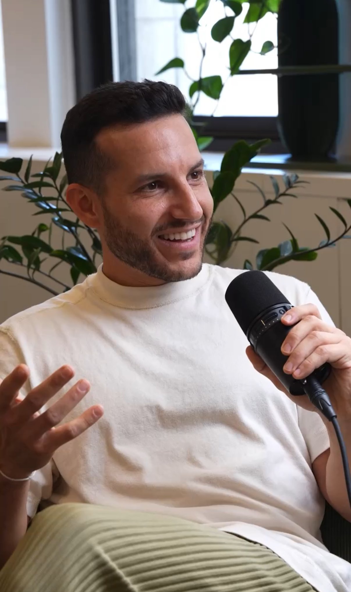 A man holding a microphone and smiling while talking, sitting indoors near a window with plants in the background.