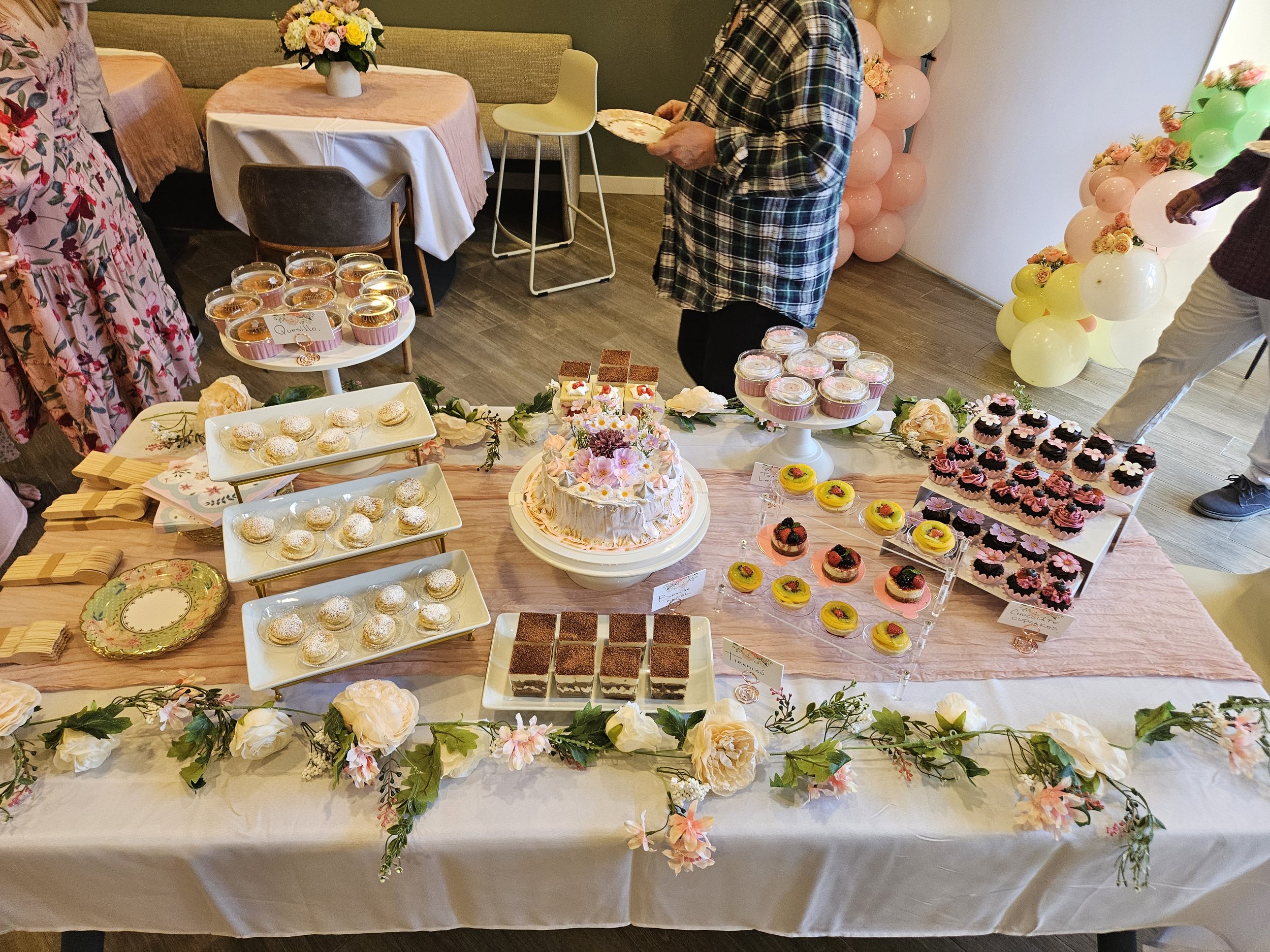 A dessert table with various sweets including cupcakes, cakes, and pastries, decorated with flowers and balloons in the background, at a celebration or party.