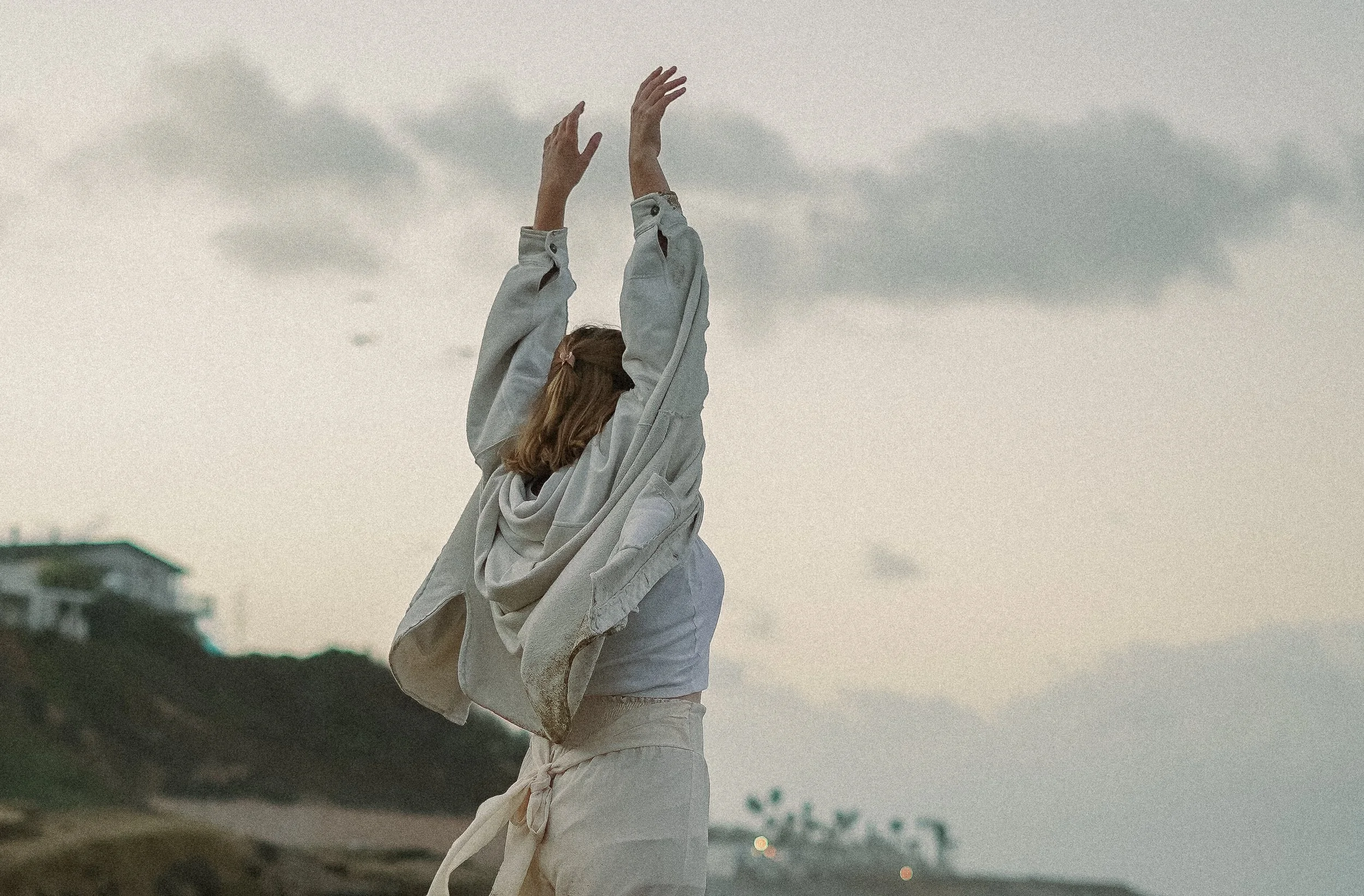 A woman in casual clothing stands outdoors with her arms raised upward, facing away from the camera, in a landscape with cloudy sky and distant buildings on a hill.