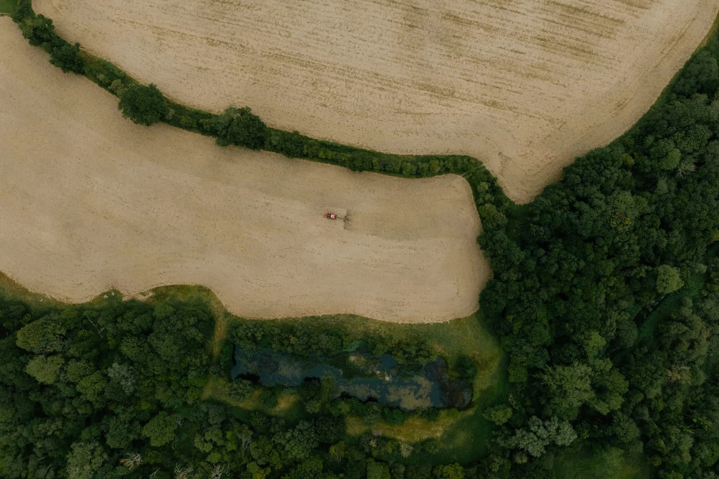 A woman with curly dark hair sitting on a bench in a room with sunlight casting shadows, wearing a beige sleeveless top and wide-legged brown pants, with a wooden paneled wall behind her.