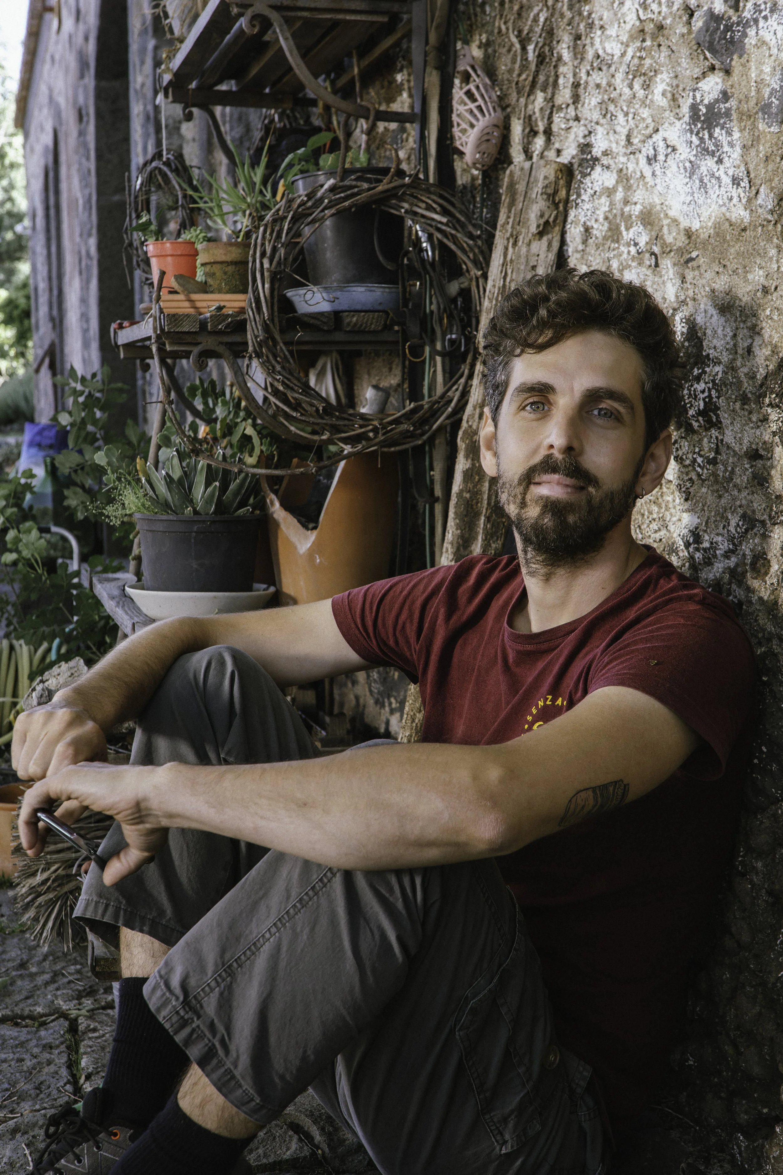 Uomo seduto con maglietta rossa, barba e capelli ricci, seduto accanto a un muro di pietra, circondato da piante e oggetti da giardinaggio.