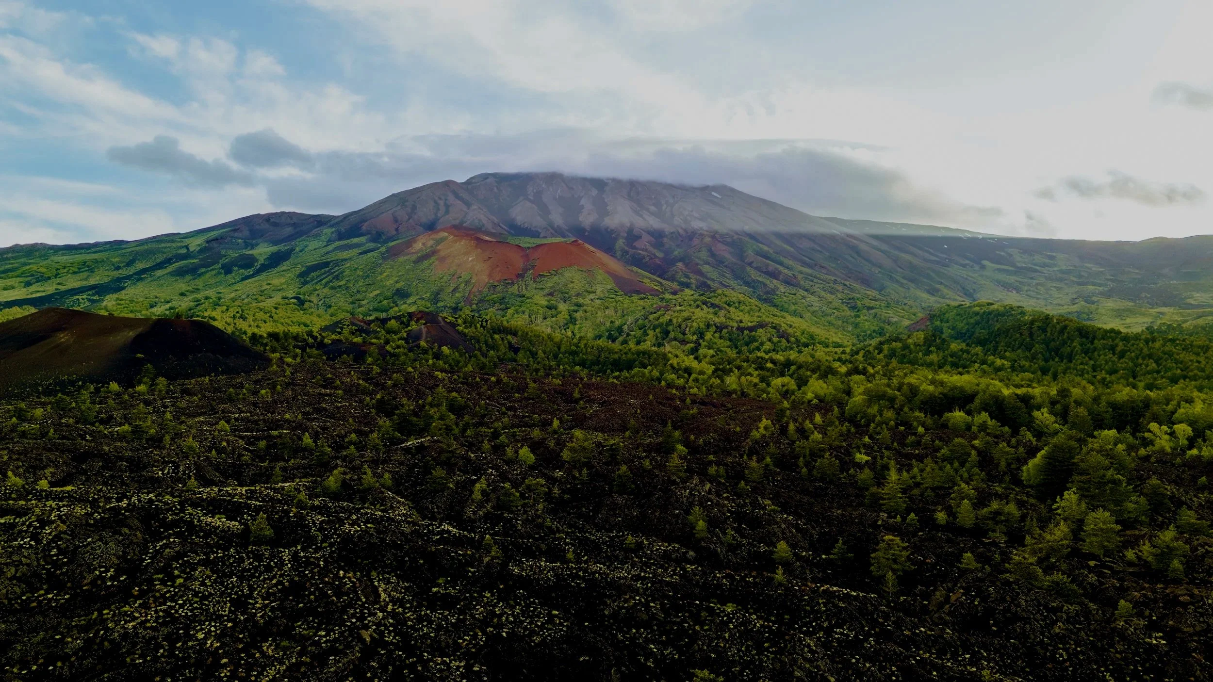 Vista di un vulcano con pendii verdi e aree di rocce scure, circondato da natura rigogliosa e cielo coperto di nuvole.