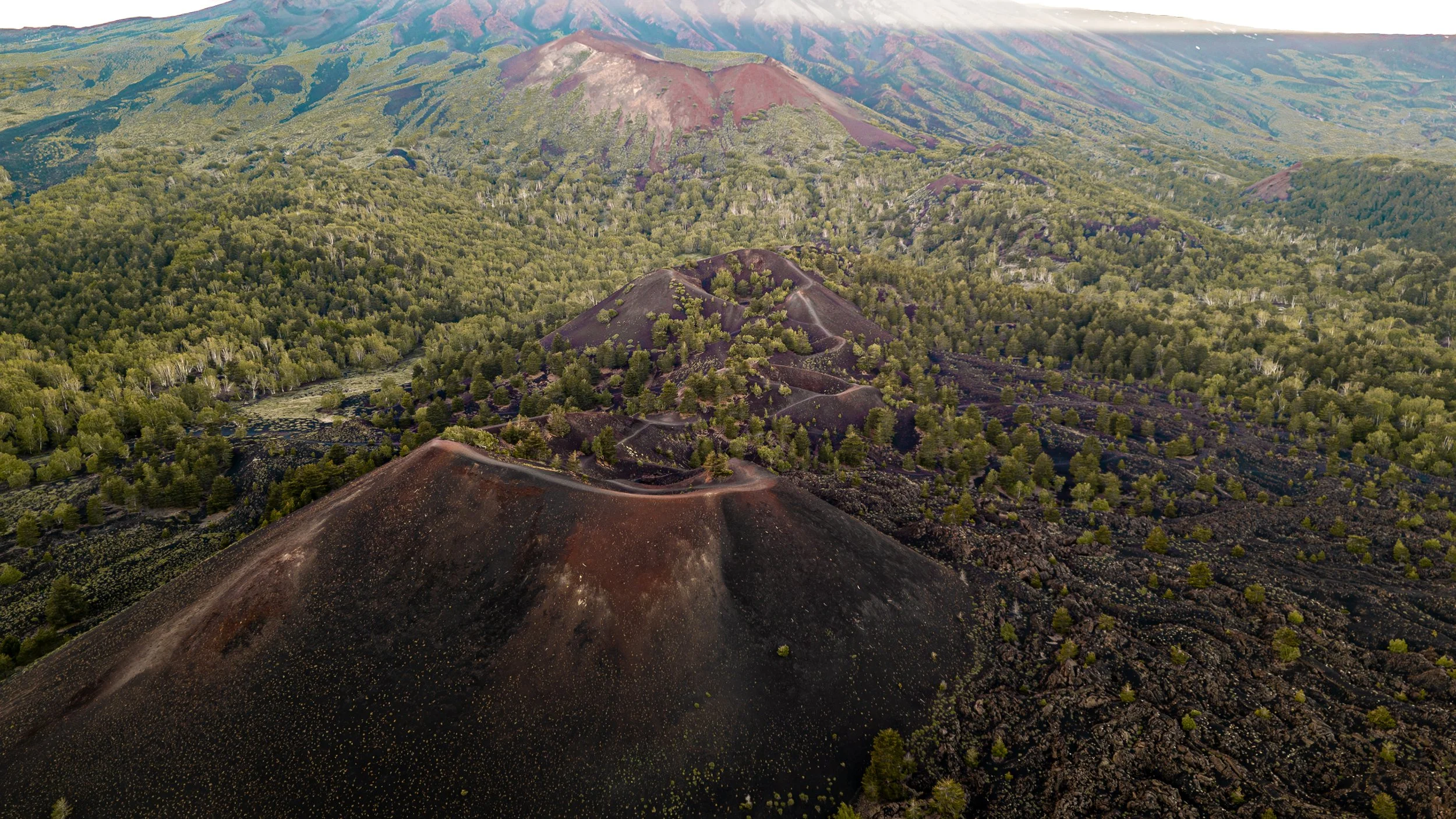 Leicota vulcanica con terreni scuri e vegetazione verde circostante, vista aerea delle colline e delle montagne in lontananza.