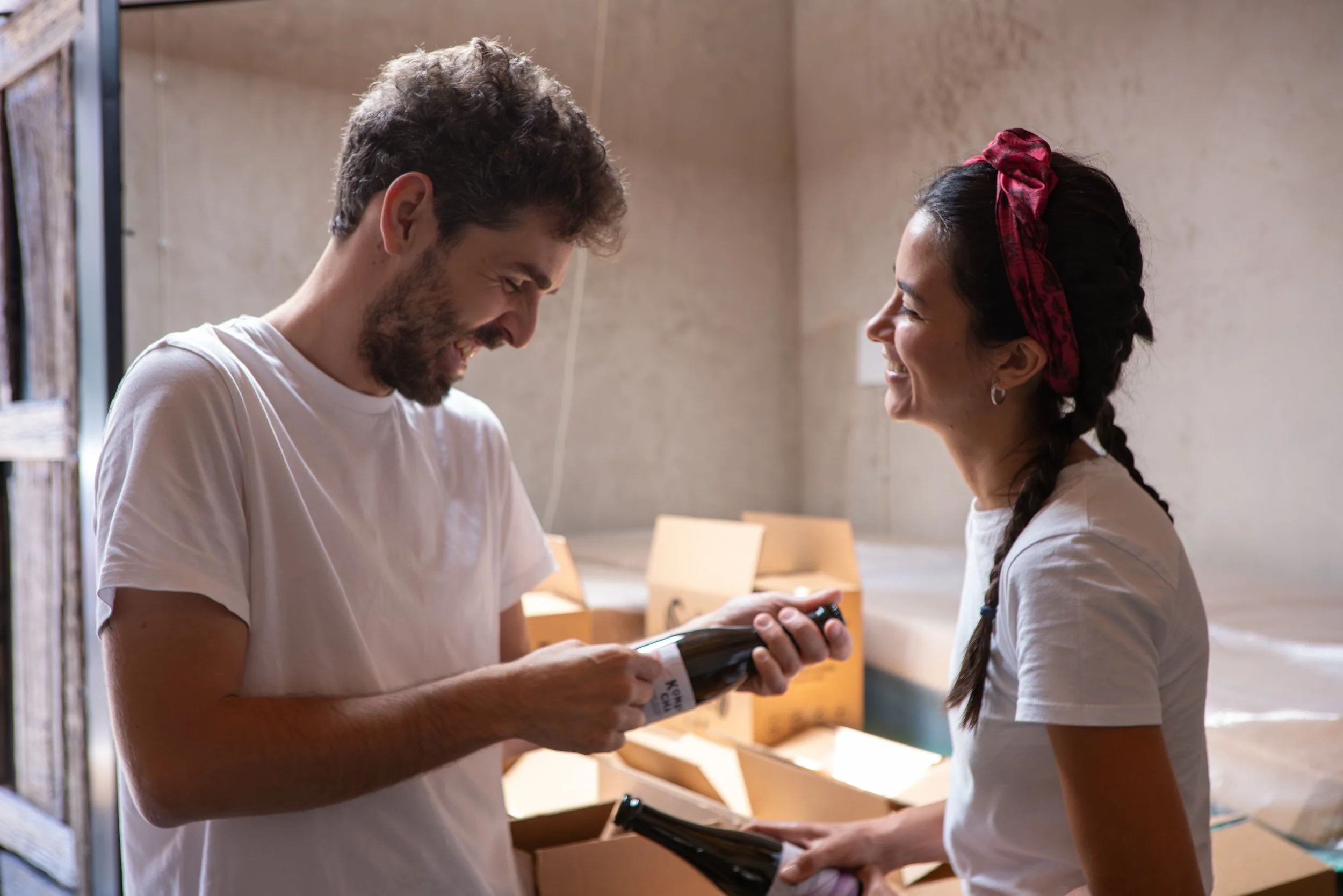 Due persone sorridenti in un ambiente industriale o di magazzino, che si scambiano bottiglie di vino, con scatole in plastica sullo sfondo.