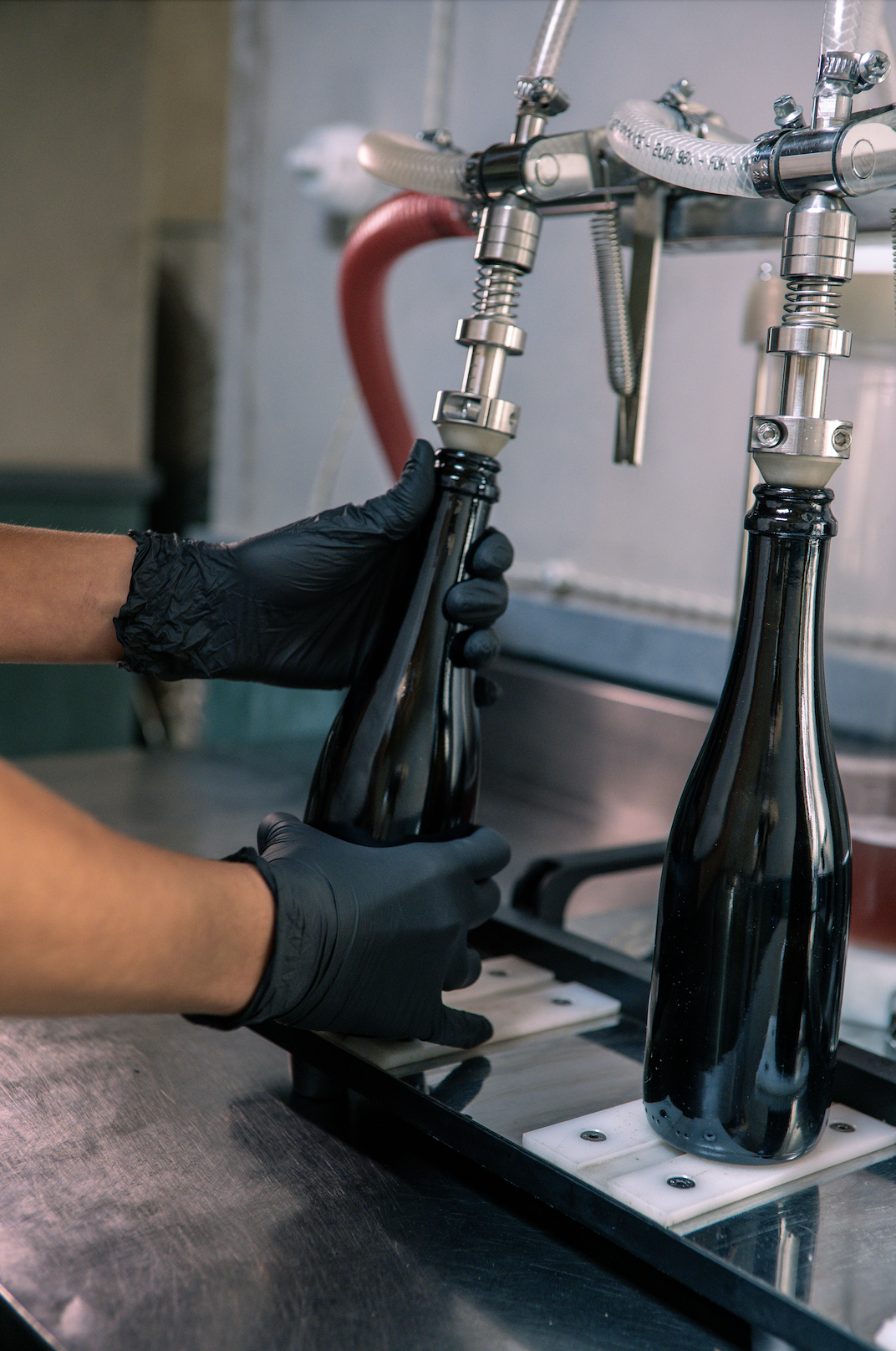 Due mani con guanti nere riempiono una bottiglia di vetro con una macchina di imbottigliamento.