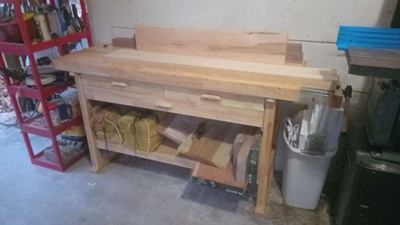 A wooden workbench in a workshop with various pieces of wood on top and beneath, a red shelving unit with tools and supplies to the left, a trash can with miscellaneous items to the right, and a gray machine in the corner.