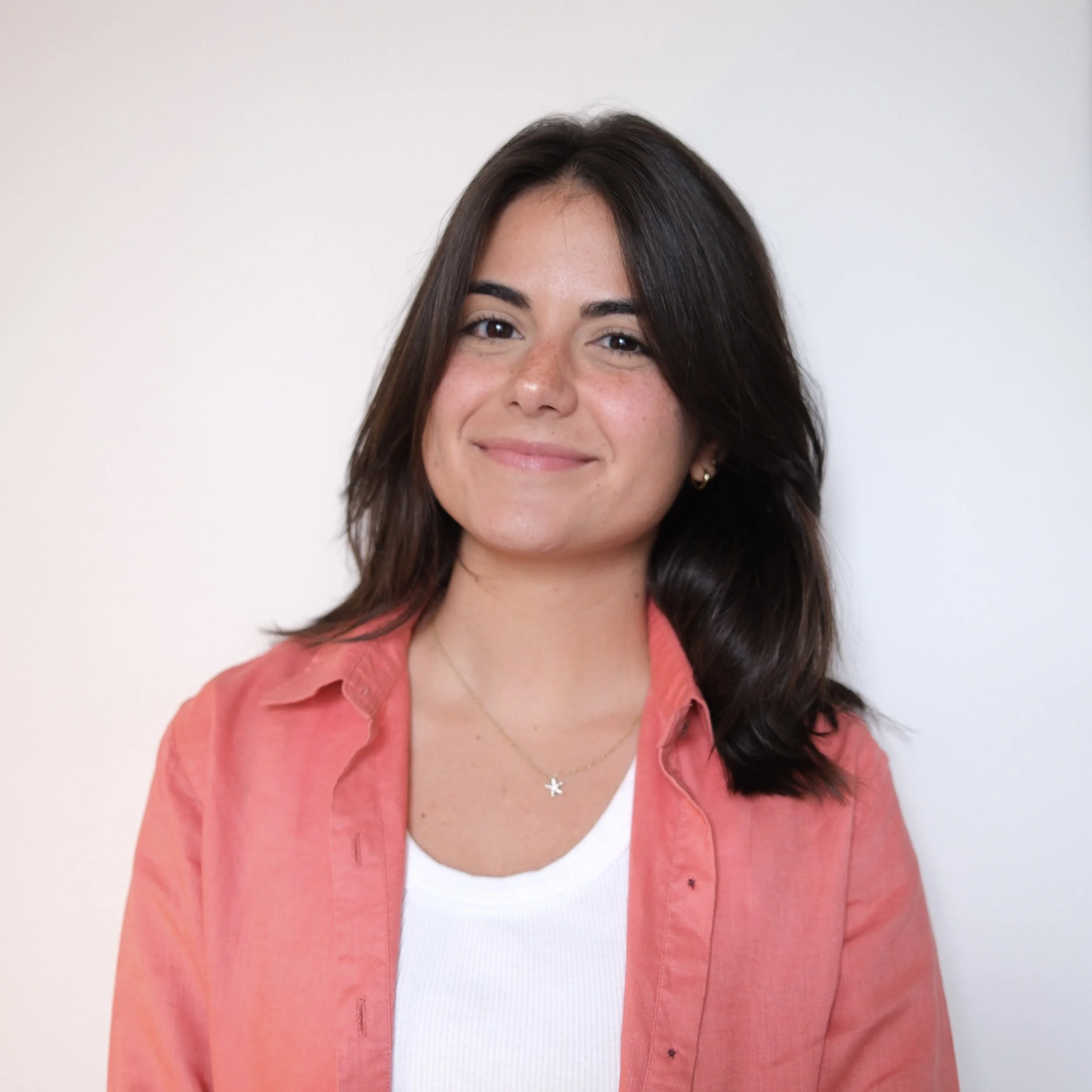 A young woman with medium-length dark brown hair, smiling, wearing a white shirt, a coral-colored jacket, small earrings, and a star-shaped necklace, standing against a plain white wall.