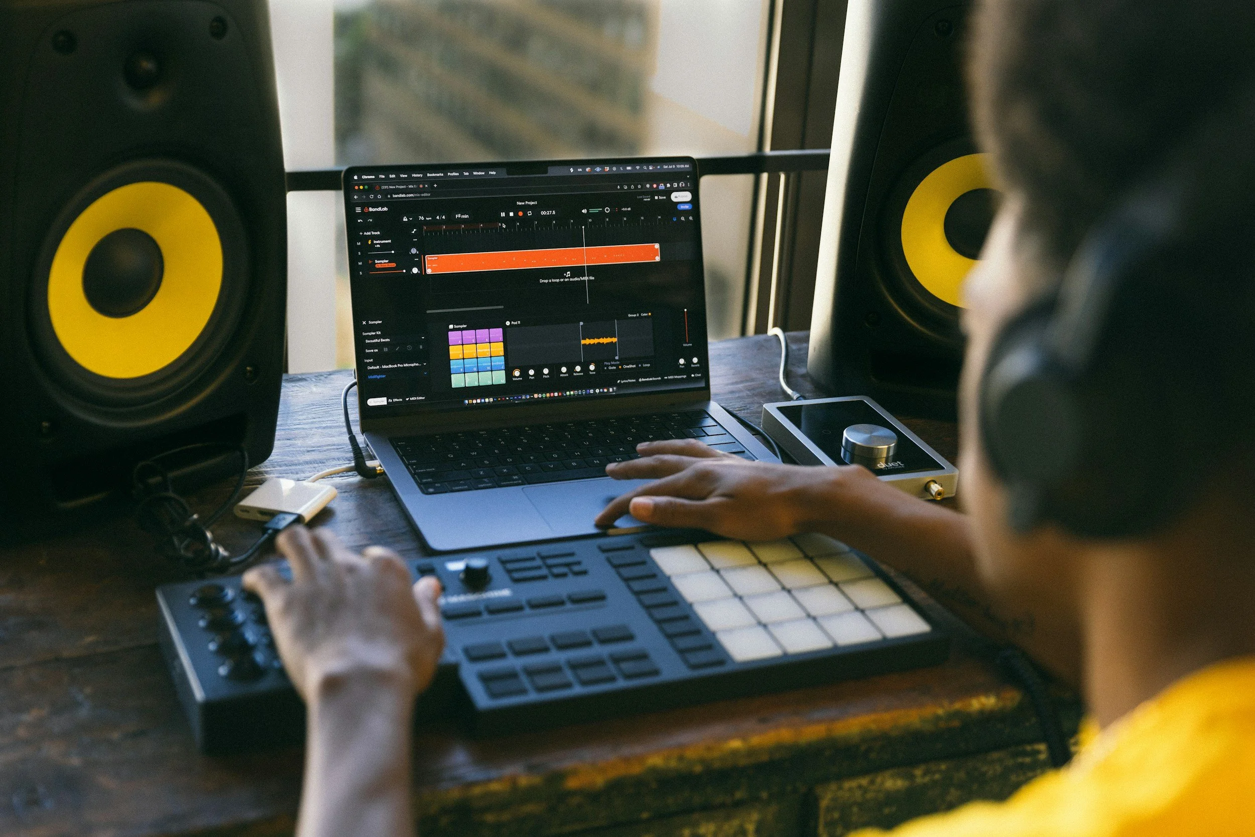 A person wearing headphones working on music production using a laptop, MIDI controller, and audio mixer, with two large yellow and black studio monitor speakers on either side.