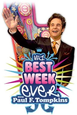 Smiling man waving next to a colorful promotional poster for 'Best Week Ever with Paul F. Tompkins,' a TV show.