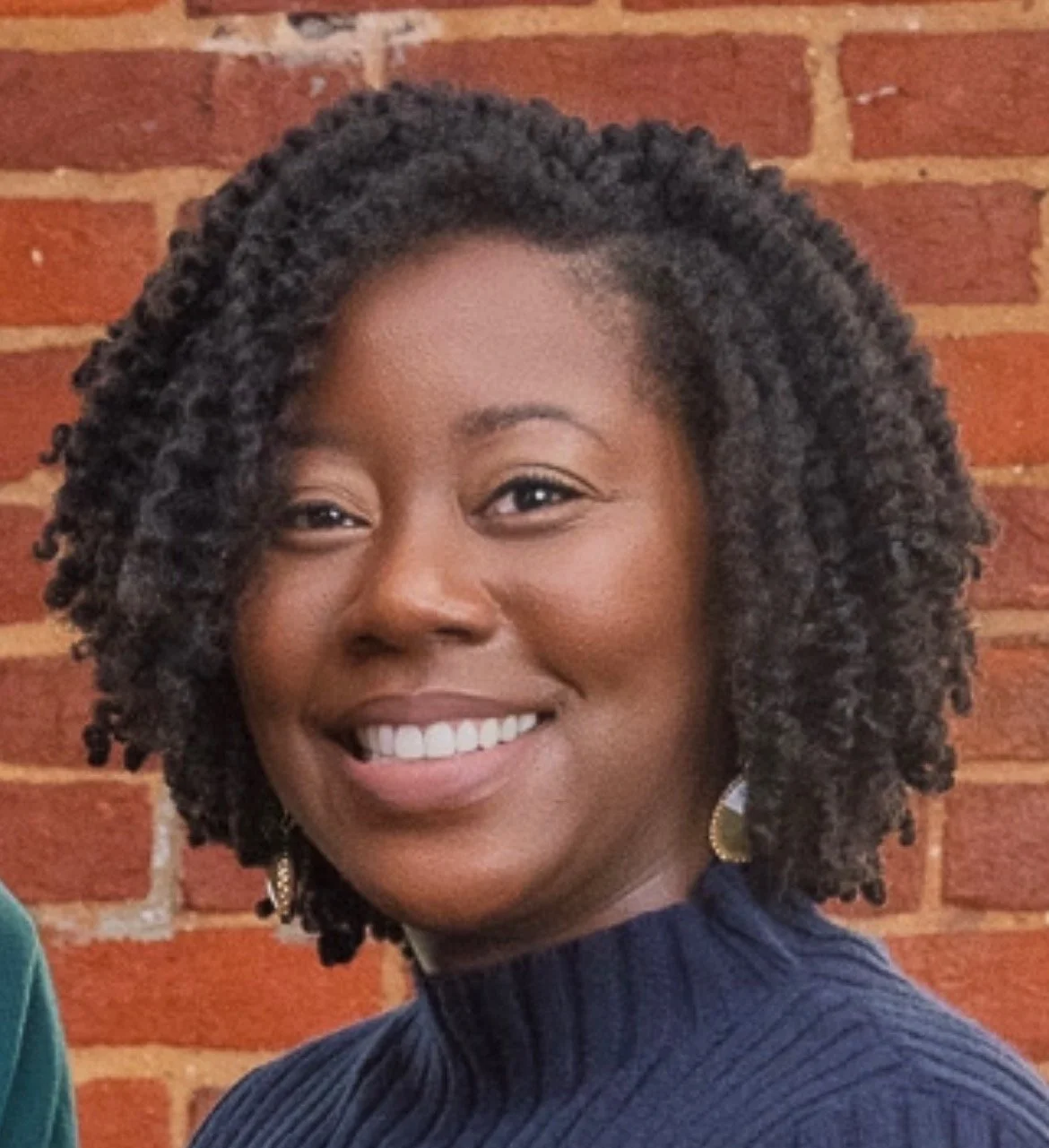 A young woman with natural curly hair, smiling, wearing a dark blue top and gold earrings, standing in front of a red brick wall.