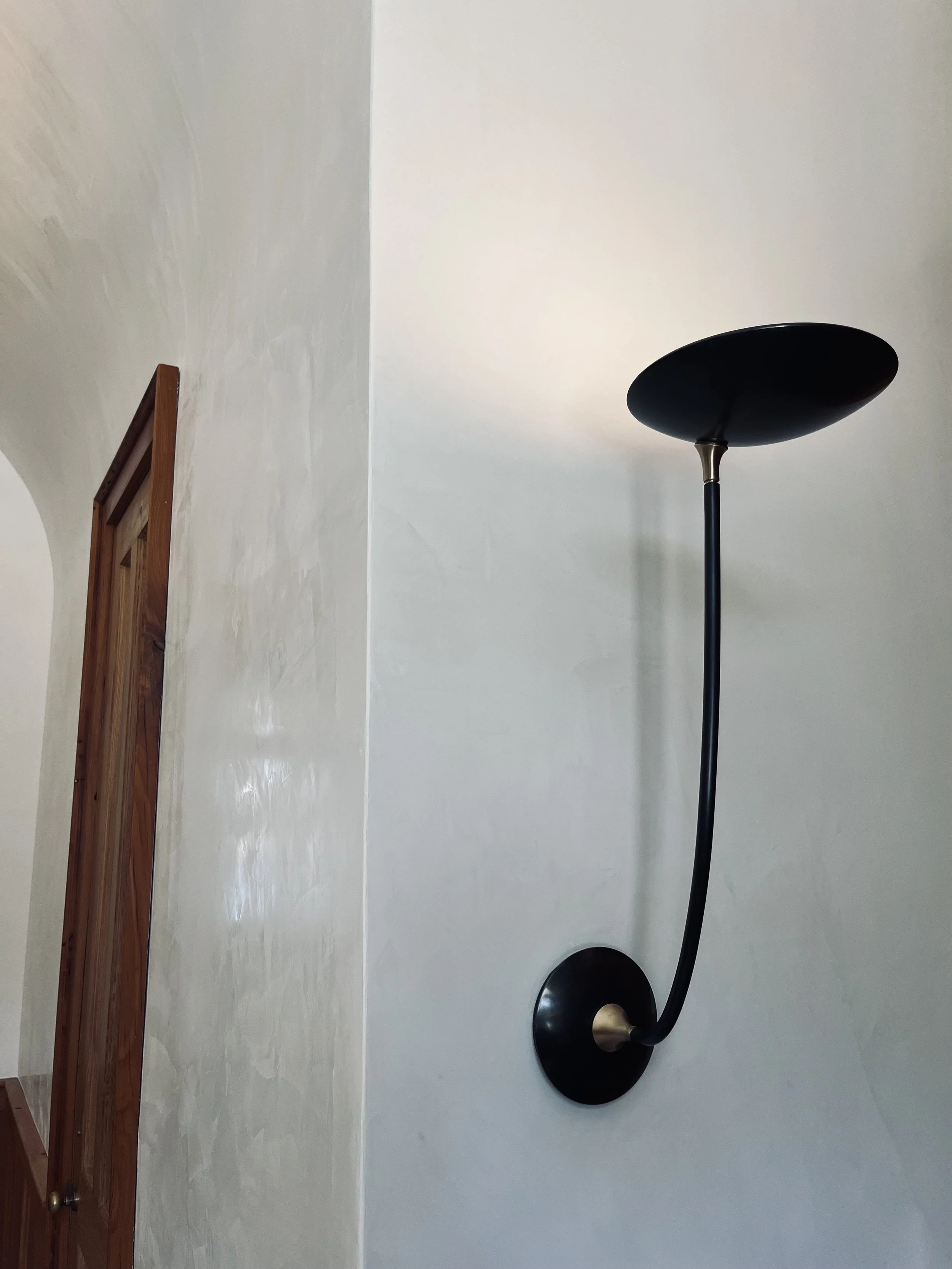 A modern wall-mounted black lamp with a flexible arm on a textured white wall inside a house.