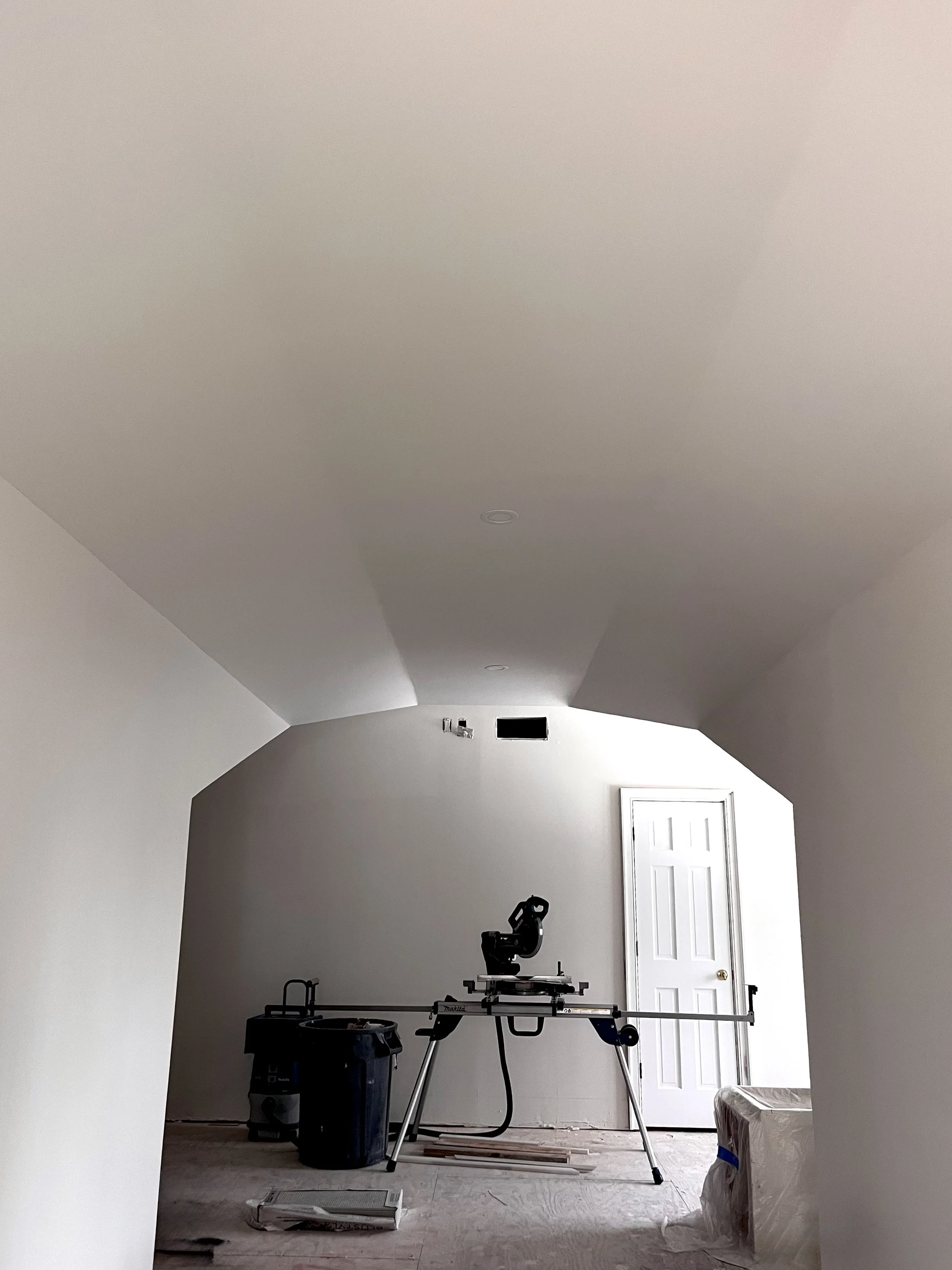 Barely furnished room with a work table, miter saw, vacuum, and a white door, possibly under renovation.