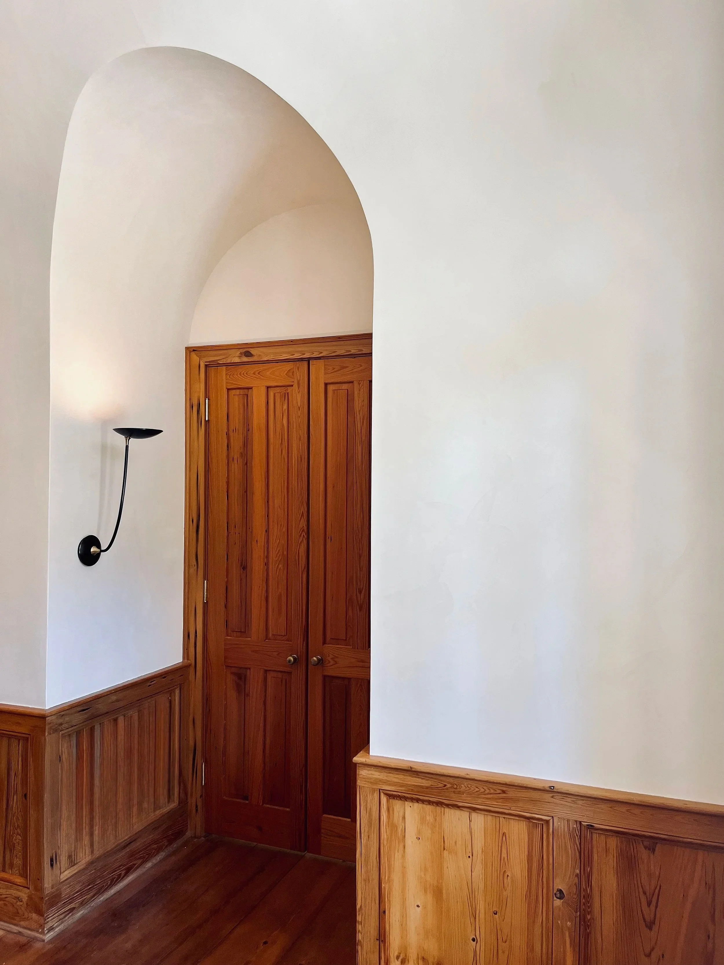 Interior view of a wooden-paneled hallway with a closed wooden door, a curved white ceiling arch, and a black wall-mounted light fixture.