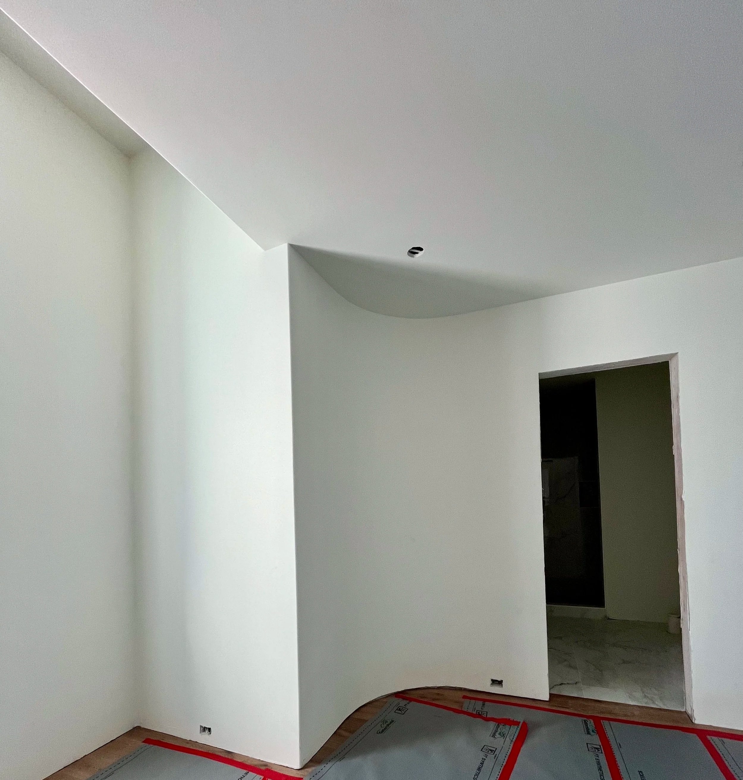 Interior of a room under construction with white walls and a curved ceiling. There is a doorway leading to another room, and construction materials are visible on the floor.