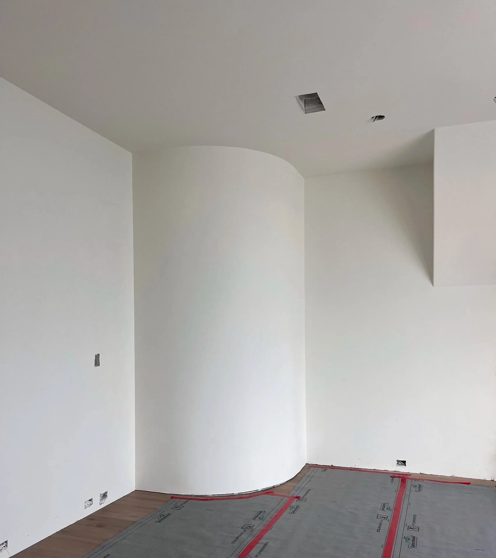 Interior of a room under construction with white walls, ceiling with vents, and protective floor covering.
