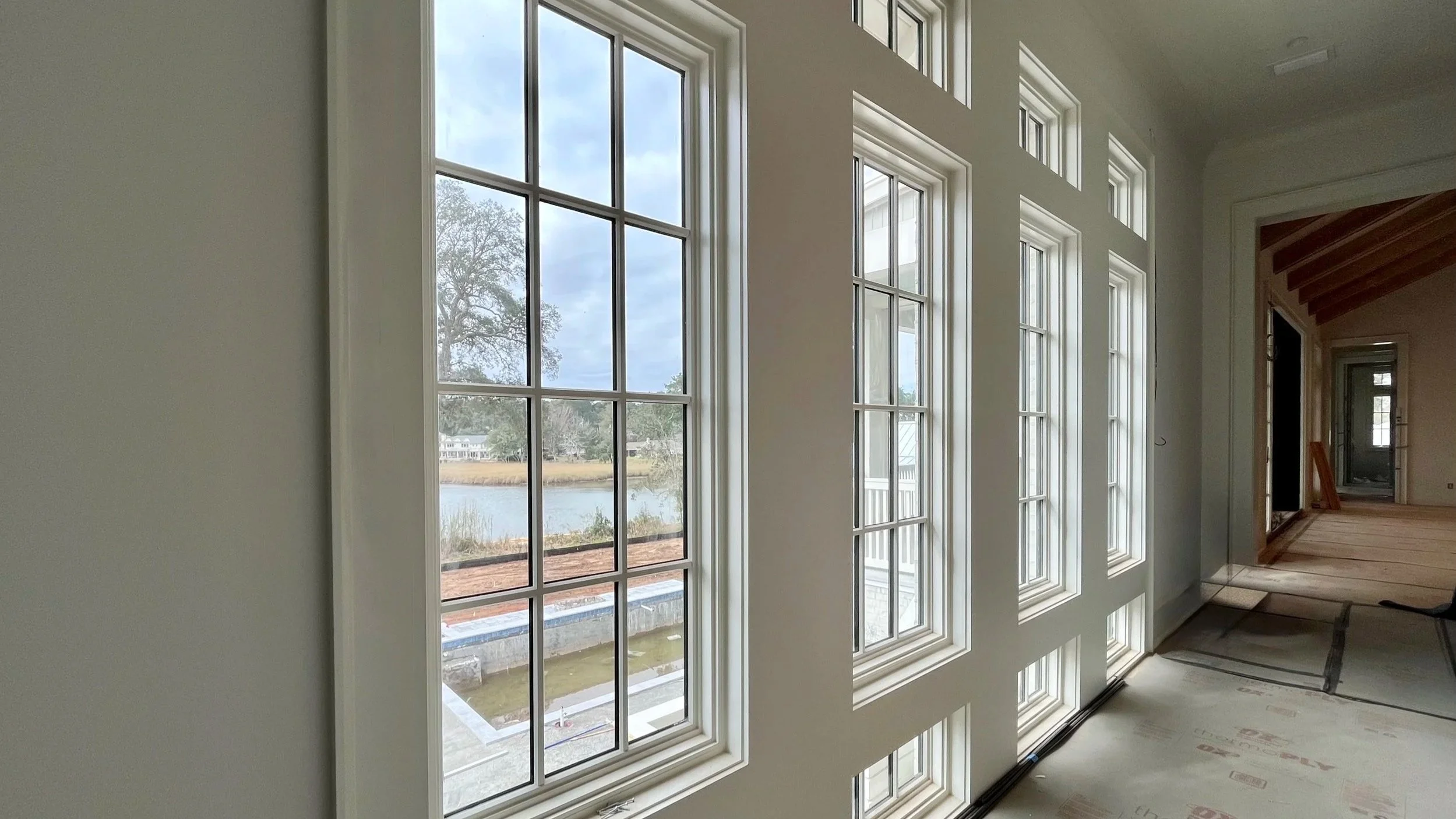 Interior of a house under construction with large windows overlooking a pond and outdoors, showing wooden flooring and partially finished walls.