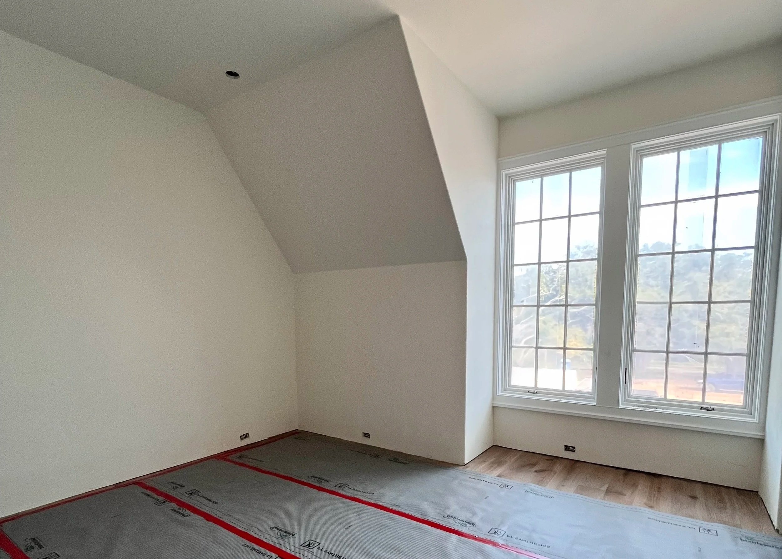 Empty room with large double window, unfinished floor with protective covering, white walls, sloped ceiling, and no furniture.