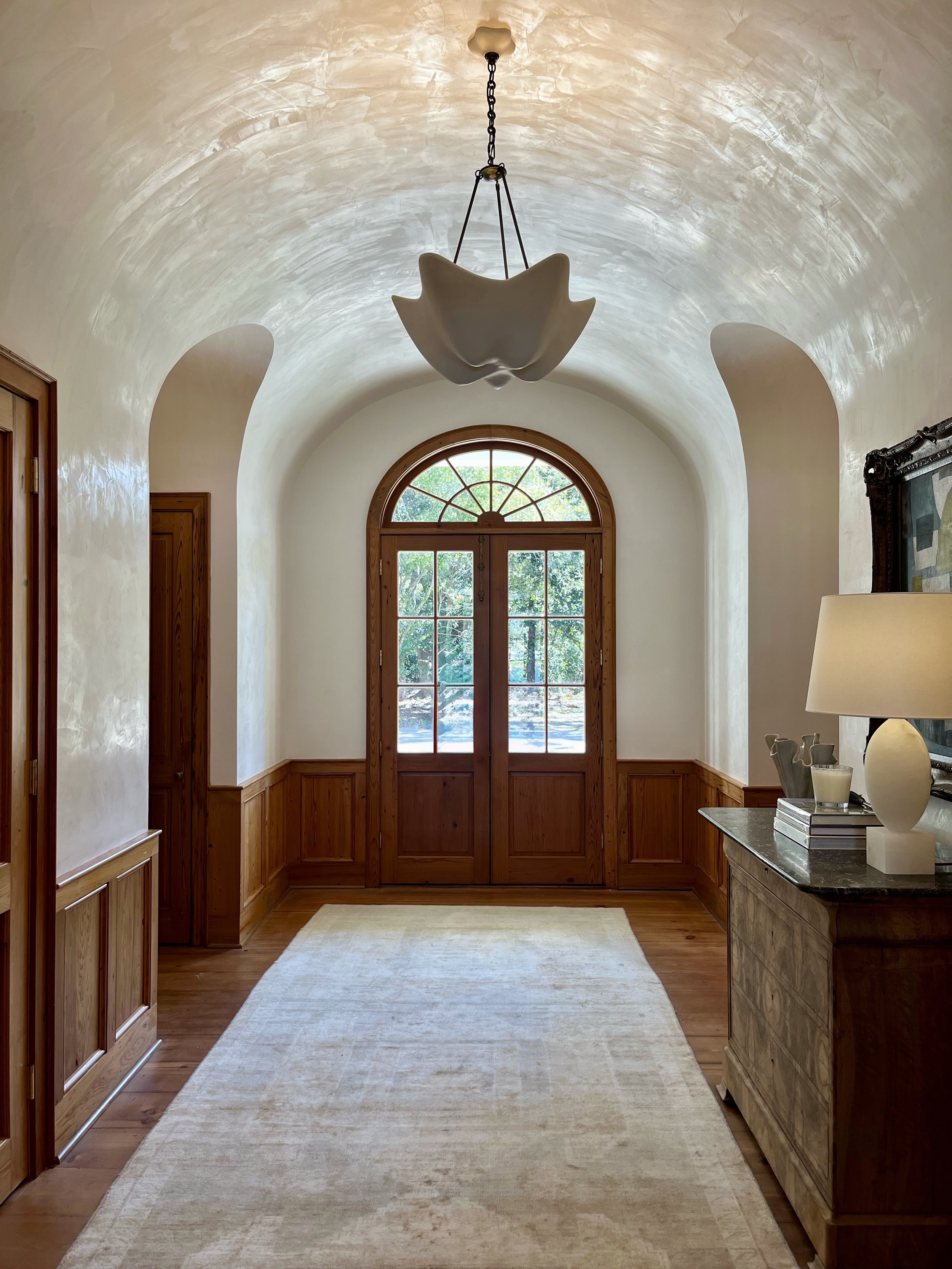 A hallway with arched ceiling and wooden double doors with a decorative semi-circular window above, a cream-colored rug on a hardwood floor, and a side table with a lamp and decorative objects.