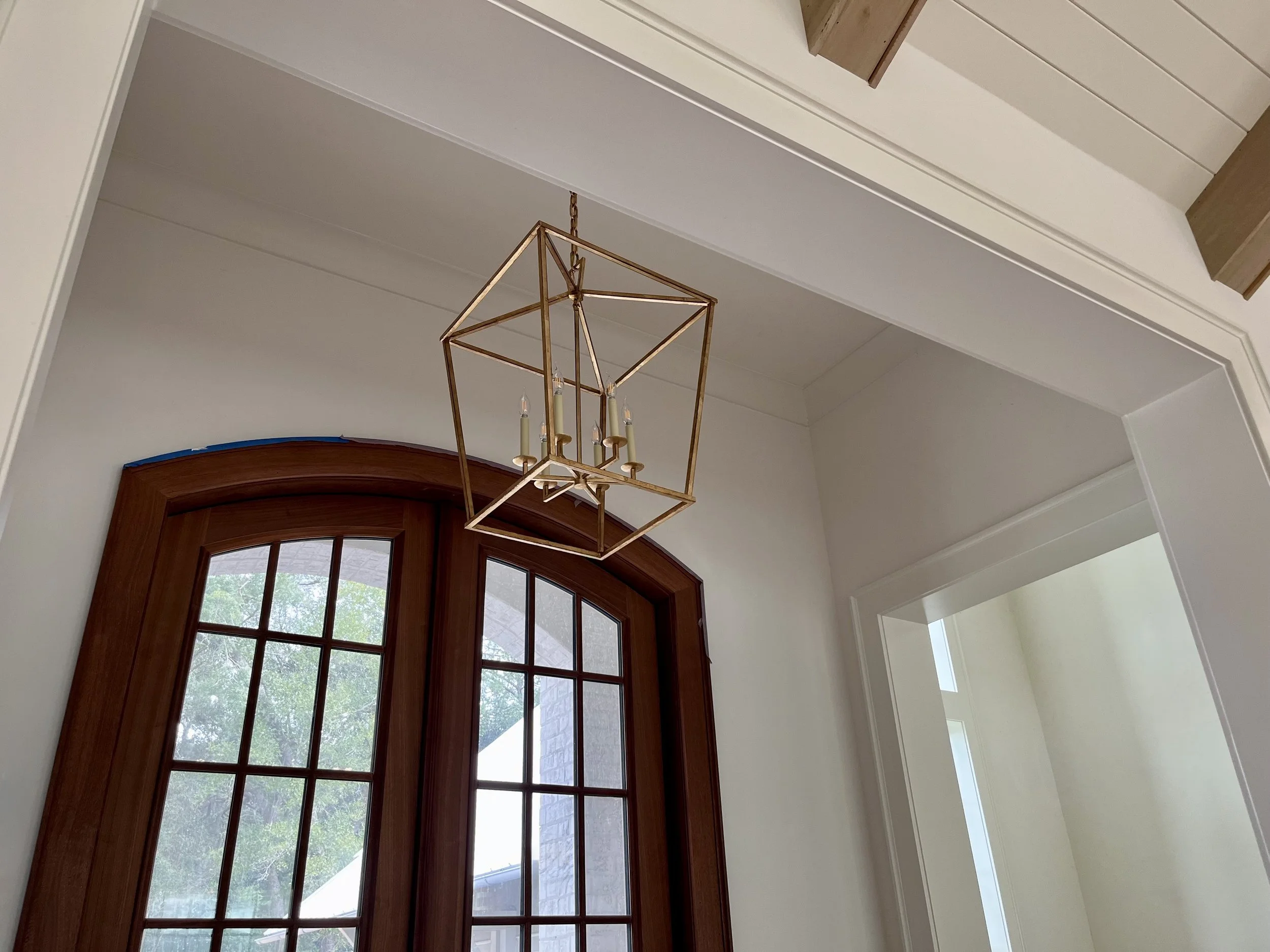 A modern chandelier with a gold geometric frame and four candle-style light bulbs hangs from the ceiling in front of a large arched window with wooden framing, revealing a green tree outside.