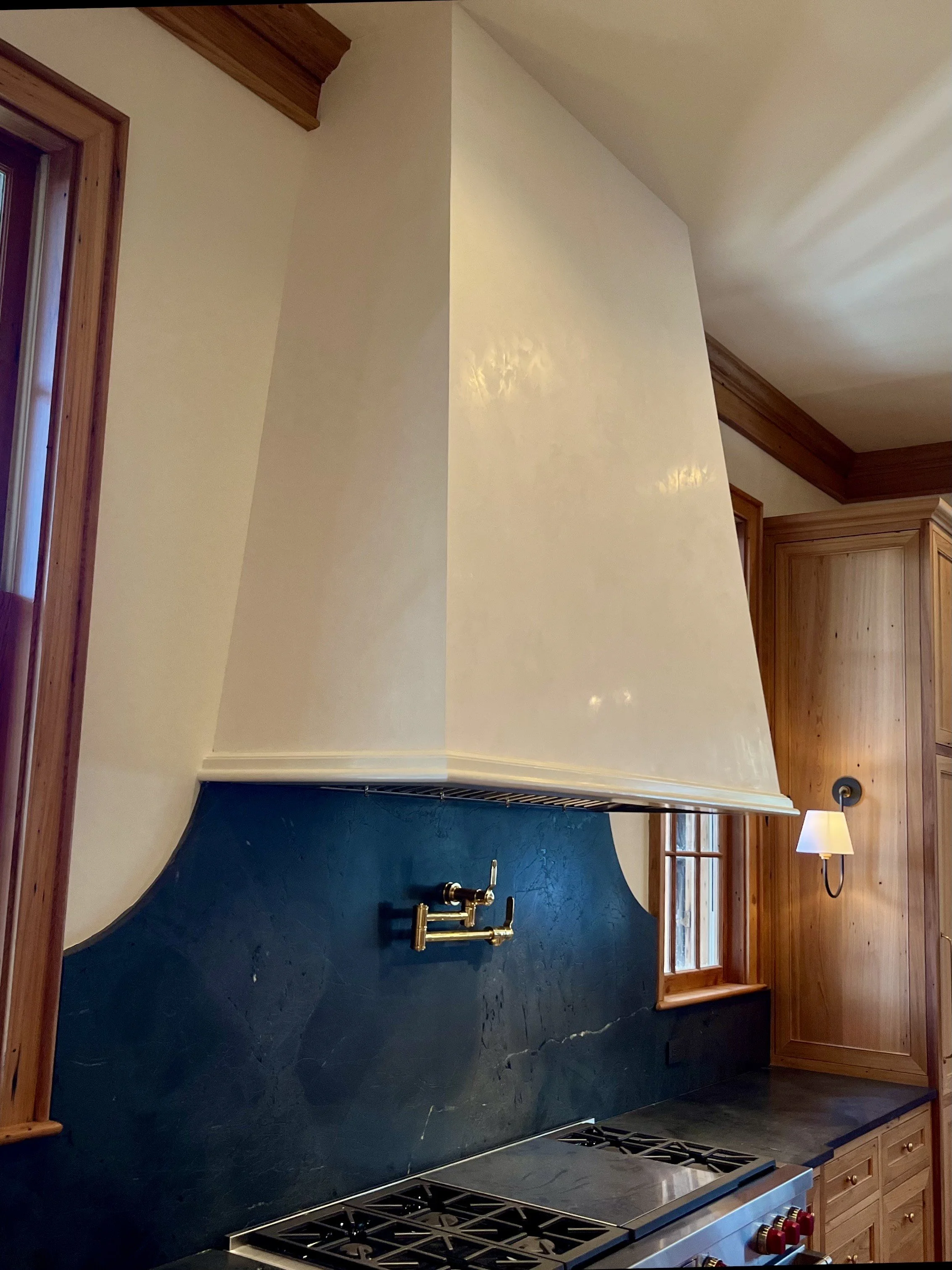 Kitchen with a large white range hood, dark green marble backsplash, wooden cabinetry, and a brass faucet above the stove.